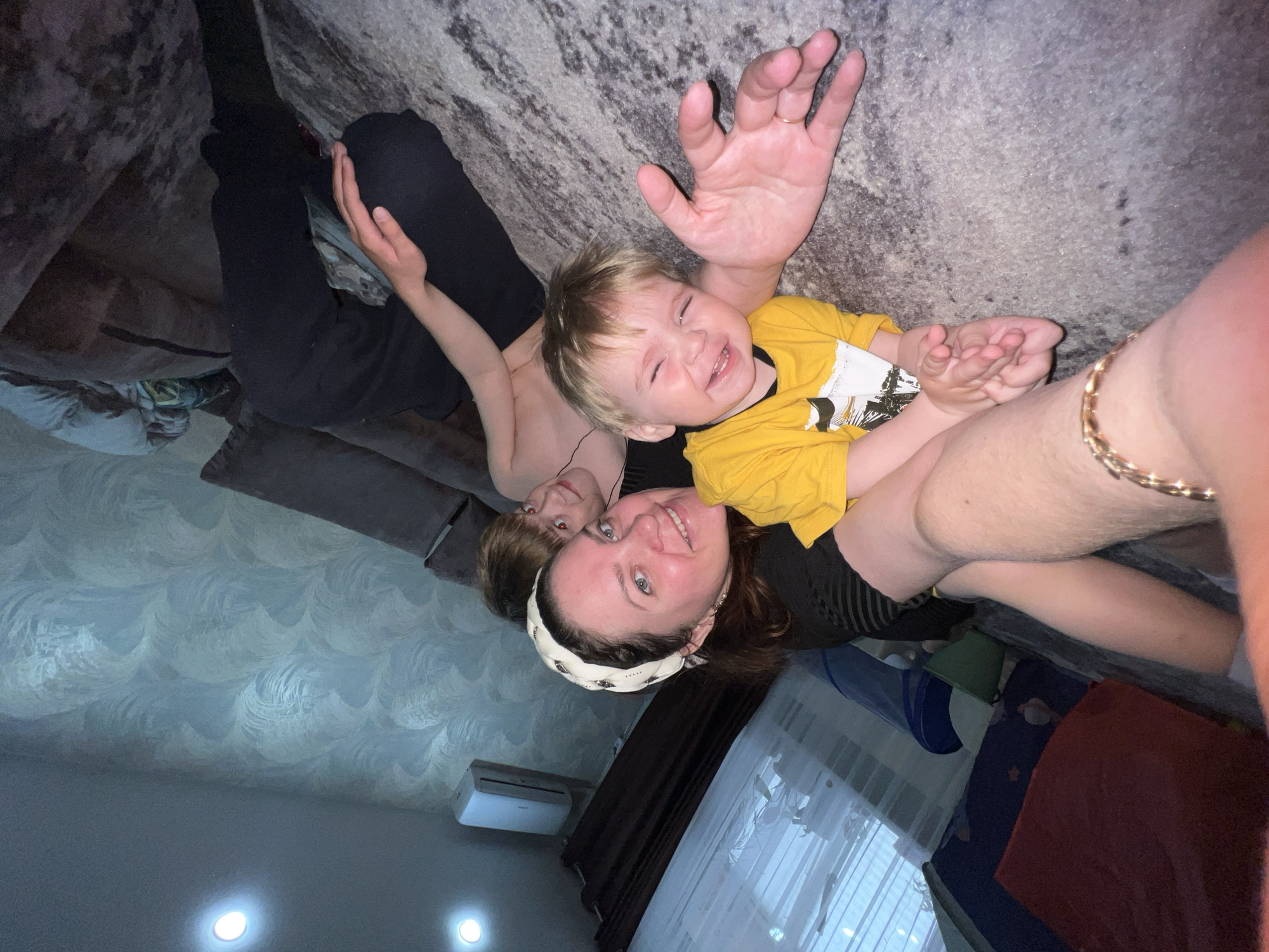 Three people are lying on a gray patterned sofa. A woman with a white headband is smiling. A young child in a yellow shirt is smiling and holding up his hands. A second person wearing black pants is also lying on the sofa. The room has textured blue walls, and there is an air conditioner near the ceiling. The location is Noviy Urengoy, Russia.