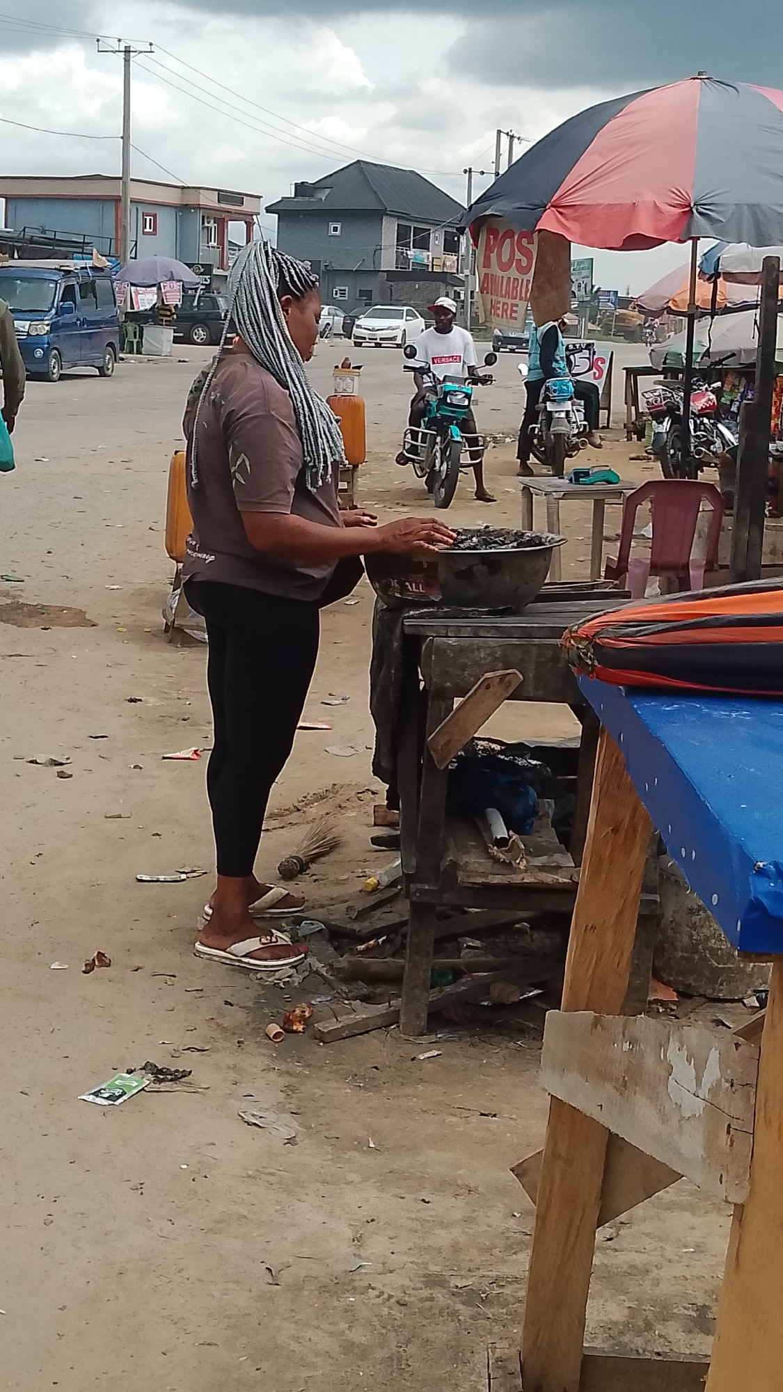 A woman with braided hair is standing in a street market in Warri, Nigeria. She is wearing a brown t-shirt and black leggings, and she is interacting with a bowl of food or goods on a wooden stand. In the background, there are several motorcycles, people on motorcycles, buildings, and street vendors with umbrellas and stalls. The ground is unpaved and littered with debris. The sky is overcast.
