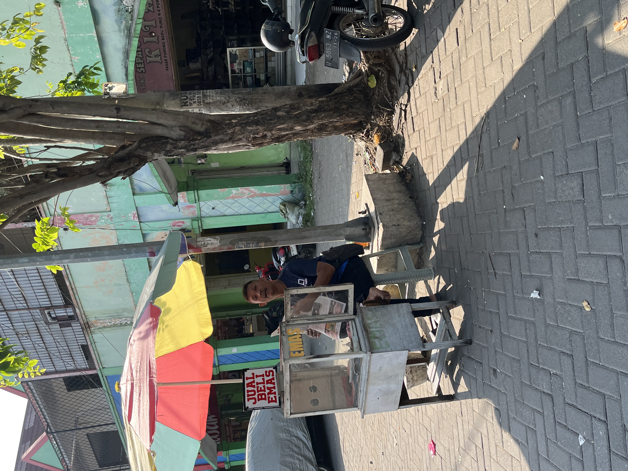 The image depicts a street scene in Juwet Kenongo, Indonesia, on a sunny day. In the foreground, a man sits on a stool next to a mobile cart, which appears to be a jewelry or pawn shop stall. A sign above the cart reads "JUAL BELI EMAS," meaning "Buy Sell Gold." The cart has a glass display case, and the man is holding what looks like a newspaper or brochure. The cart is positioned on a paved sidewalk, with a tree to the right. A motorcycle is parked behind the tree, and a parked car is partially visible to the left. The background shows buildings with green and pastel facades, typical of Indonesian architecture, and foliage from trees. The overall impression is of a bustling local marketplace or commercial street.