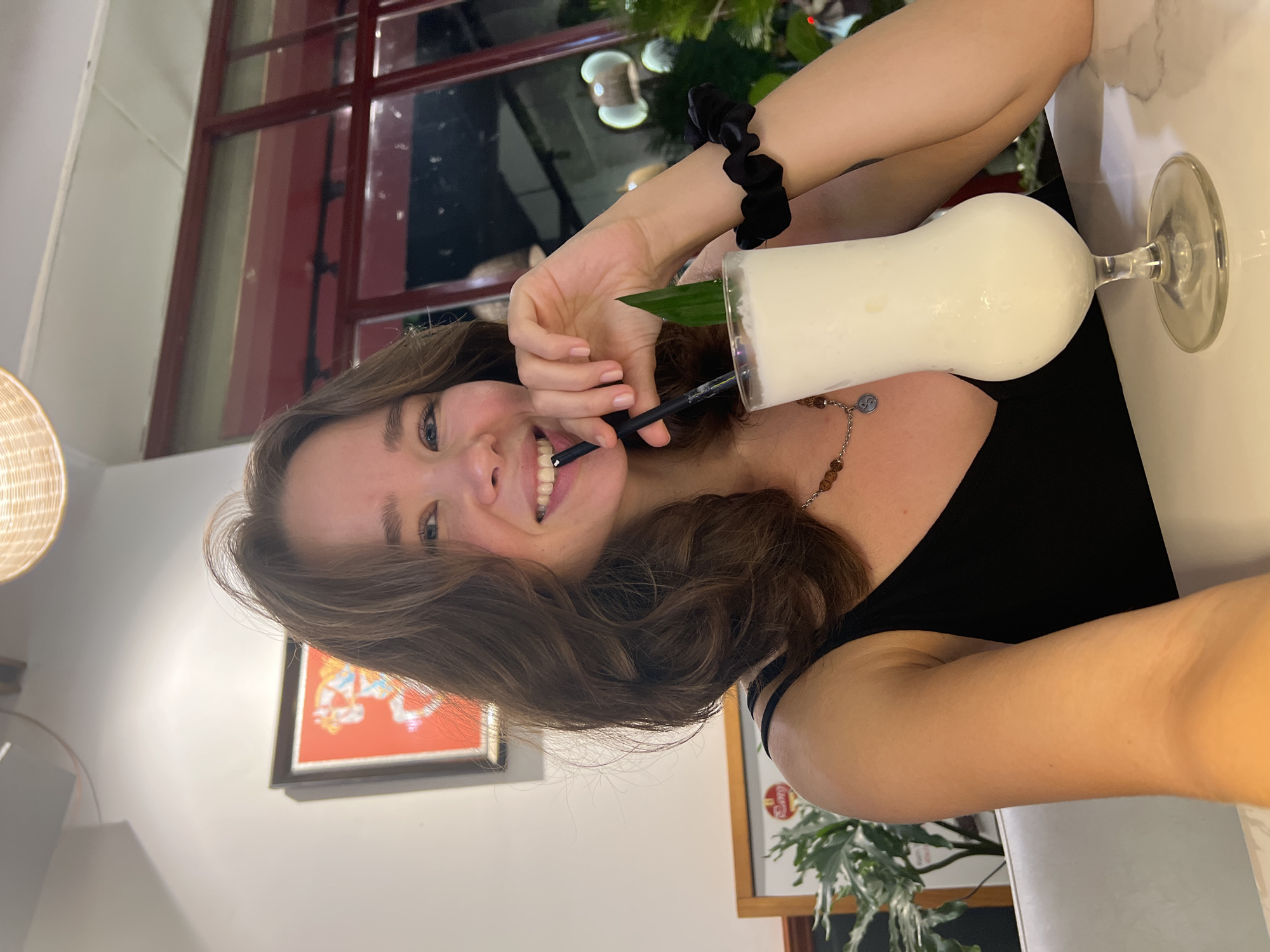 A light-skinned woman with wavy brown hair is posing for a selfie indoors, holding a cocktail in a tall glass with a straw and a green leaf. The cocktail appears to be white and creamy. The woman is wearing a black top, and a black scrunchie is visible on her wrist. She has a necklace with a pendant and bead details. She is smiling and holding the straw in her mouth.
The background includes a window with a red frame and some greenery. The location appears to be in Bắc Nha Trang, Vietnam, judging by the setting, possibly a restaurant or cafe with local decor. Part of a framed picture on the wall is also visible. A woven lampshade hangs out of frame on the left.