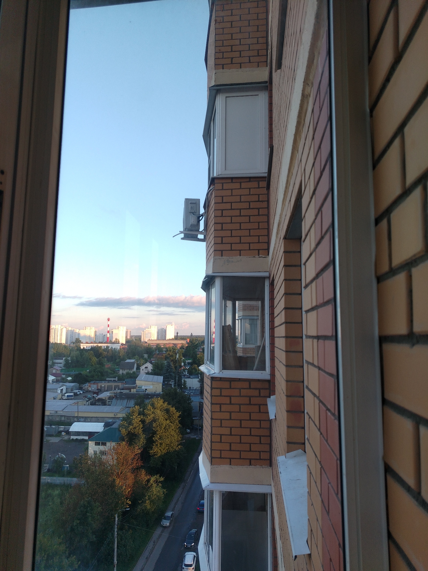 The image is a vertical, slightly angled shot taken from an elevated position, likely a balcony, looking outwards and downwards. The foreground is dominated by the edge of a window frame on the left and the brick facade of a building on the right. The building facade is constructed with reddish-brown and lighter tan bricks. Visible on the building's exterior are window frames, some with white casements, and an air conditioning unit mounted on a bracket.
The background reveals a city skyline under a clear sky with a few wispy clouds, suggesting either early morning or late afternoon. A cluster of multi-story apartment buildings forms the prominent feature of the cityscape, interspersed with trees and smaller structures. A tall, industrial chimney with red and white banding is also visible in the distance. Below the apartment buildings, a street is visible with a few parked cars and moving vehicles. The presence of residential buildings, apartment complexes, and industrial elements suggests an urban or suburban environment.
The location of Andreyevka, Russia, is not explicitly indicated by any visual cues within the image itself. The description provided here is based solely on the visual content of the photograph.
