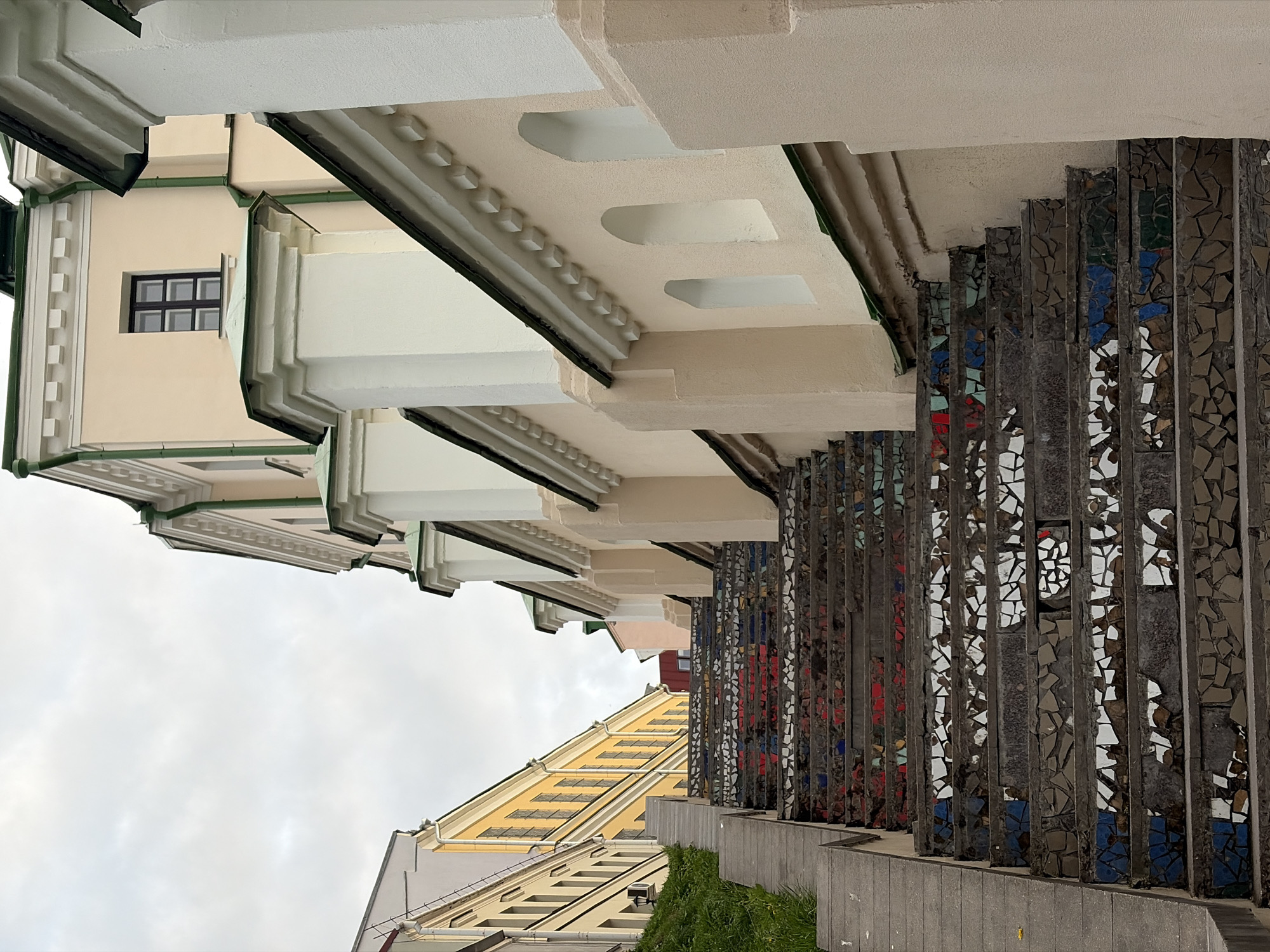 The image shows an upward view of buildings and a decorative stairway in an urban area. The buildings feature alternating levels and architectural details, including window patterns, trim, and cornices. The stairway, with mosaic decorations, creates vertical lines that contrast with the horizontal lines of the buildings. The sky is overcast. The location is unknown, as this information is not available in the image.