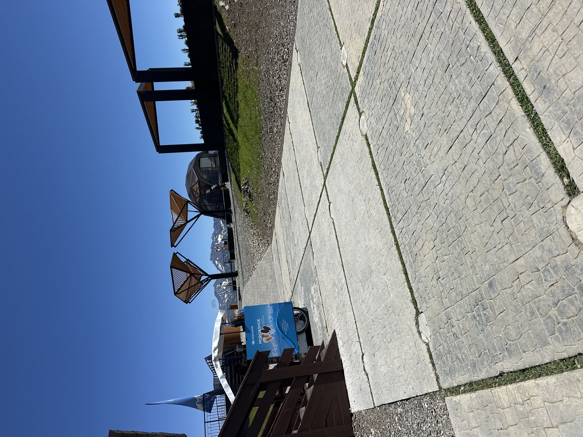 The image depicts an outdoor scene with a clear blue sky and snow-capped mountains in the background. In the foreground, a paved walkway made of textured concrete slabs with grass growing between them leads towards a structure with a domed roof and open-air seating areas with slatted roofs. To the left, a wooden staircase descends, and a blue ice cream cart is visible with Russian text indicating "ROSA KHUTOR" and "MOROZHENOE" (ice cream). A tall blue flag with a white emblem is also present on the left side of the image. The environment appears to be a mountain resort area, likely in Estosadok, Russia, given the "ROSA KHUTOR" branding. The scene is static and captures a bright, sunny day.