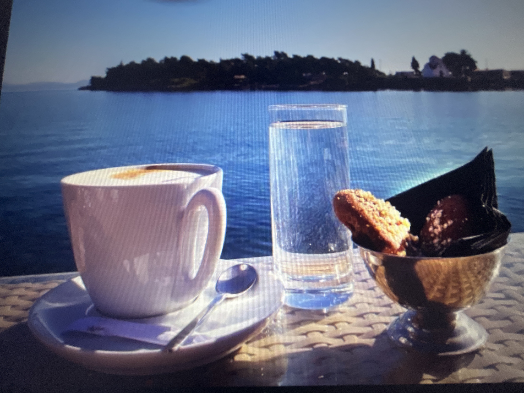 A close-up, high-angle, slightly blurry photograph shows a breakfast setting on a woven textured table in front of a body of water. The setting includes a white ceramic cup and saucer with a coffee inside, a small silver spoon on the saucer, and a sugar packet. To the right of the cup is a tall glass of clear liquid, possibly water. Beside the glass is a small metallic dish containing two small pastries or candies, one topped with nuts and the other with dark seeds, presented in a black paper wrapper. In the background, a silhouetted island covered in dark foliage with some structures on it is visible across the blue water, under a clear, pale blue sky. The location is specified as Varna, Bulgaria. No people are visible in the image.