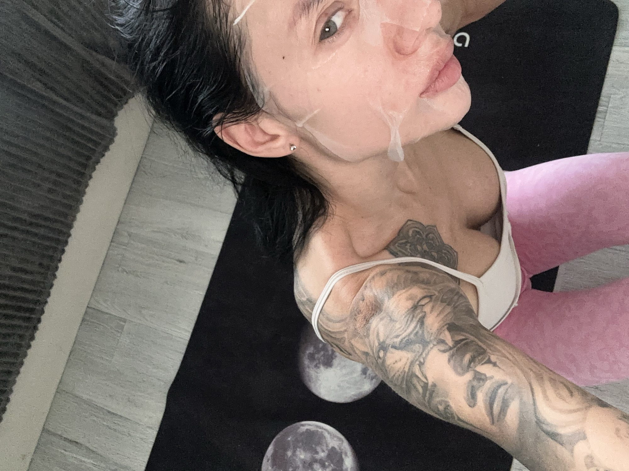 A close-up, high-angle shot shows a woman in a white sports bra and pink leggings. She has a sheet mask on her face and a tattoo on her left arm. She is lying on a black mat with a moon phase design. The mat is on a light-colored wooden floor. A dark grey, ribbed object is visible in the upper right corner, possibly a radiator or bed frame. The image does not contain any identifiable landmarks or elements that would place it specifically in Moscow, Russia.