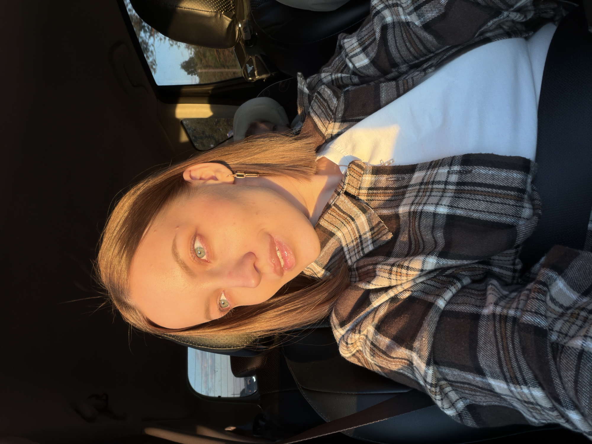 A woman is seated in a car wearing a white t-shirt and a brown and tan plaid flannel shirt. She is wearing a seatbelt. She has blonde hair and a gold geometric earring in her left ear. The scene is illuminated by warm, golden sunlight. The location is unclear, and there is no indication of being in Rogachovo, Russia.