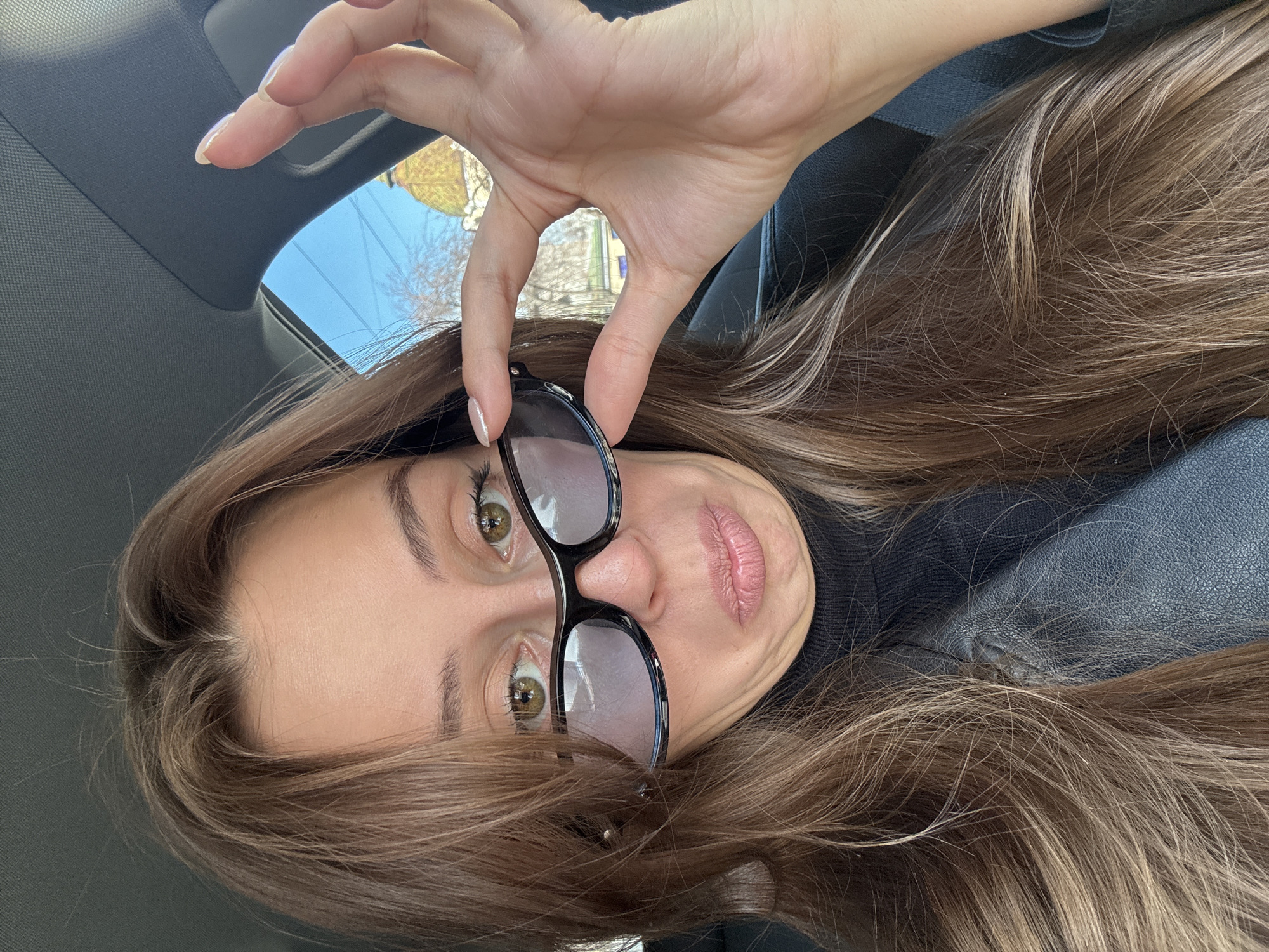 A woman is holding sunglasses in front of her face. She has long brown hair, hazel eyes, and is wearing a black turtleneck. The photo appears to be taken inside a car, with the car interior visible. The background visible through the car window suggests an urban environment. While it is not possible to verify the location in Krasnoyarsk, Russia based on the image alone, the photograph shows a woman in what looks like an urban setting.
