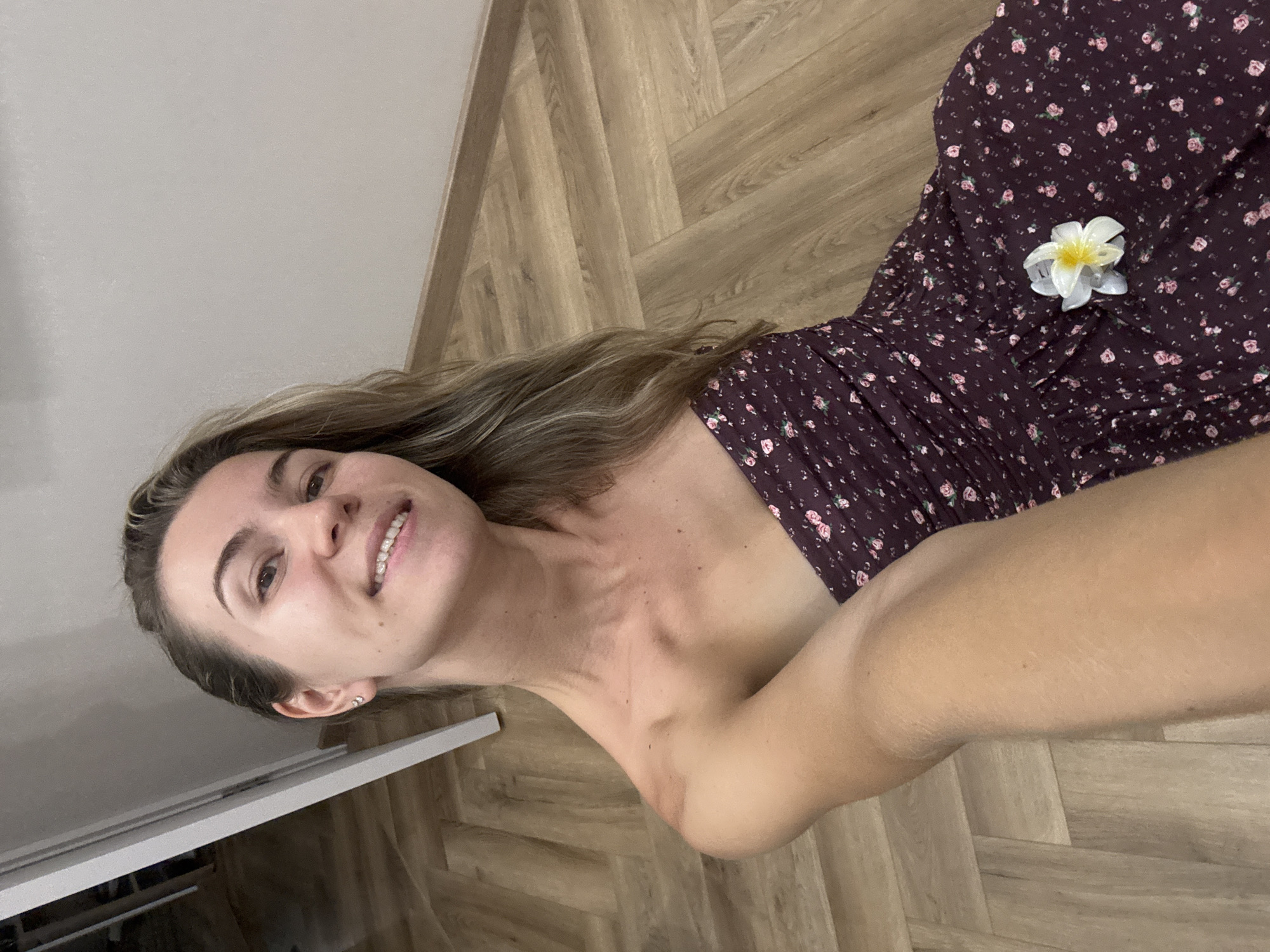 An overhead, medium shot shows a woman in a strapless, dark purple floral-print dress sitting on a light brown, wood-paneled floor. A white and yellow flower is pinned to the dress on her right side. The woman has long, wavy brown hair and is smiling at the camera, with her left arm extending forward, out of frame. She is wearing small, diamond stud earrings. The background consists of a light grey wall and a section of a white doorframe. The lighting is artificial and soft.
