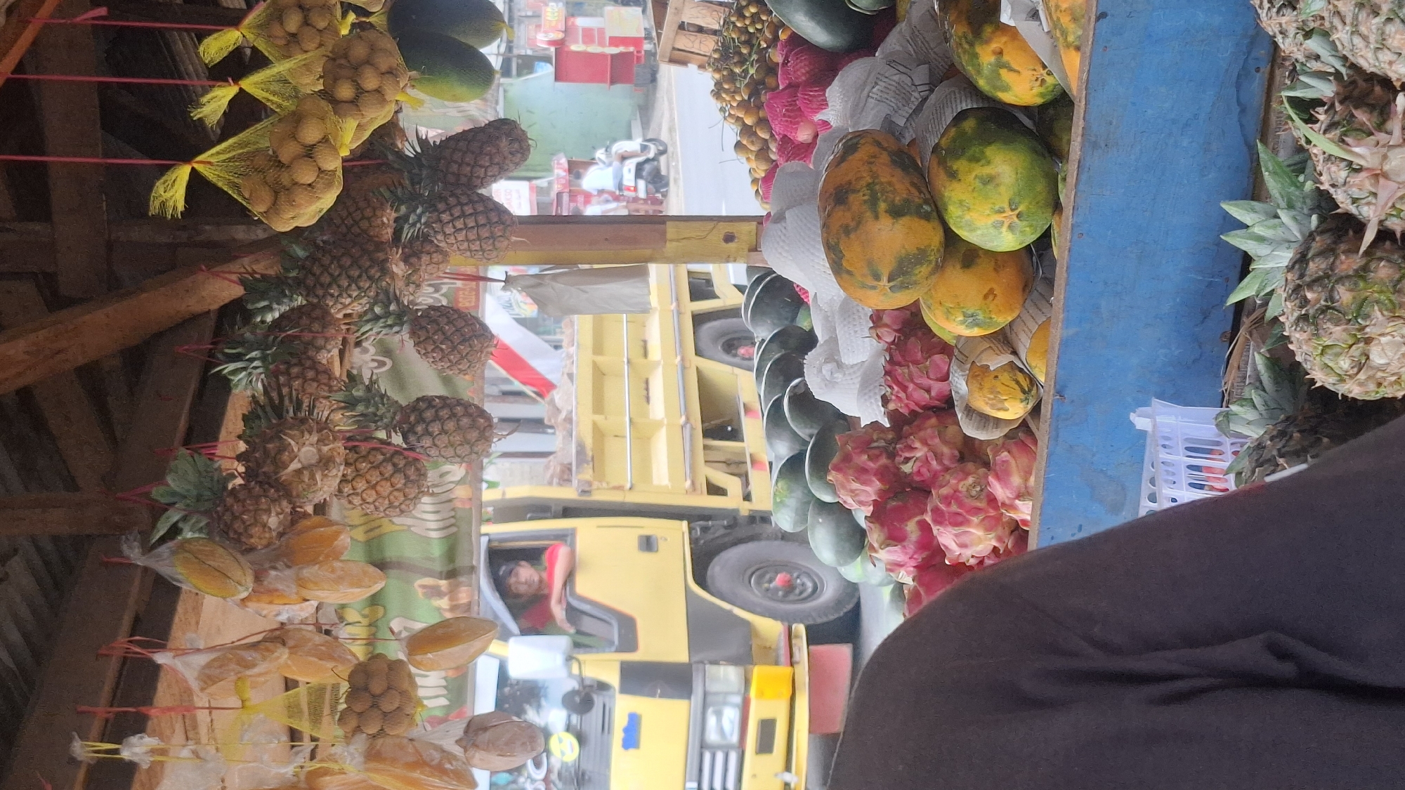In Cilaku, Indonesia, a fruit stand is set up under a wooden awning. A variety of fruits are displayed, including pineapples hanging overhead, starfruit, what appear to be lychees in yellow mesh bags, watermelons, dragon fruit, papayas, and oranges. In the background, a yellow truck is parked, with a driver visible inside. The truck has a red and white flag attached to its side. Further in the background, there are buildings, a motorcycle, and people walking. The scene depicts a typical market environment with fresh produce and the presence of vehicles and pedestrians.