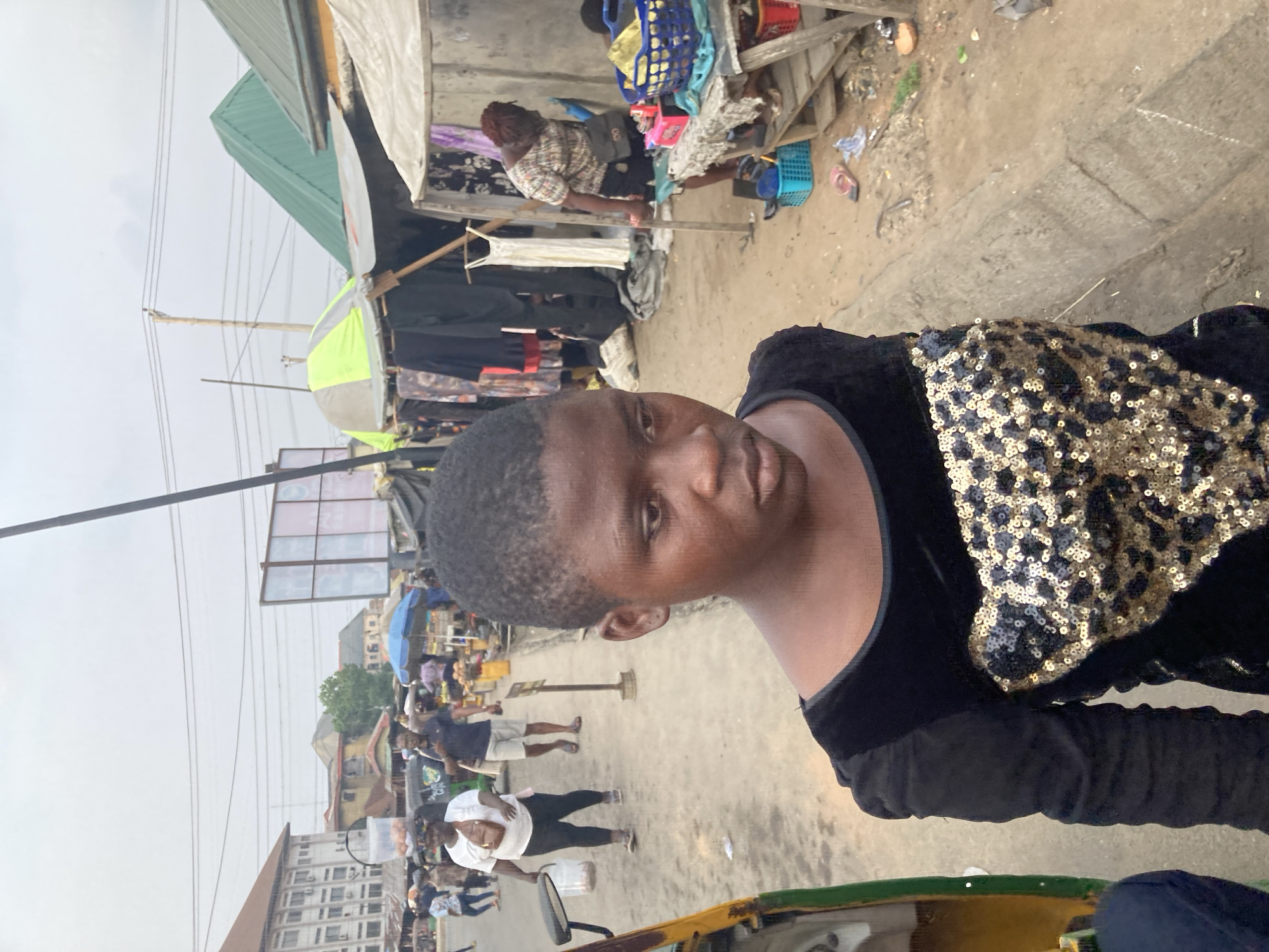The image shows a young person, possibly female, with dark hair and skin, wearing a black top with gold sequined detailing. She appears to be in Aba, Nigeria, based on the surrounding environment.
The background reveals a bustling outdoor market street scene. Numerous people are seen walking and interacting amidst vendor stalls covered with corrugated metal or cloth. There is general litter. Overhead electrical cables crisscross the sky. The lighting suggests it's daytime.