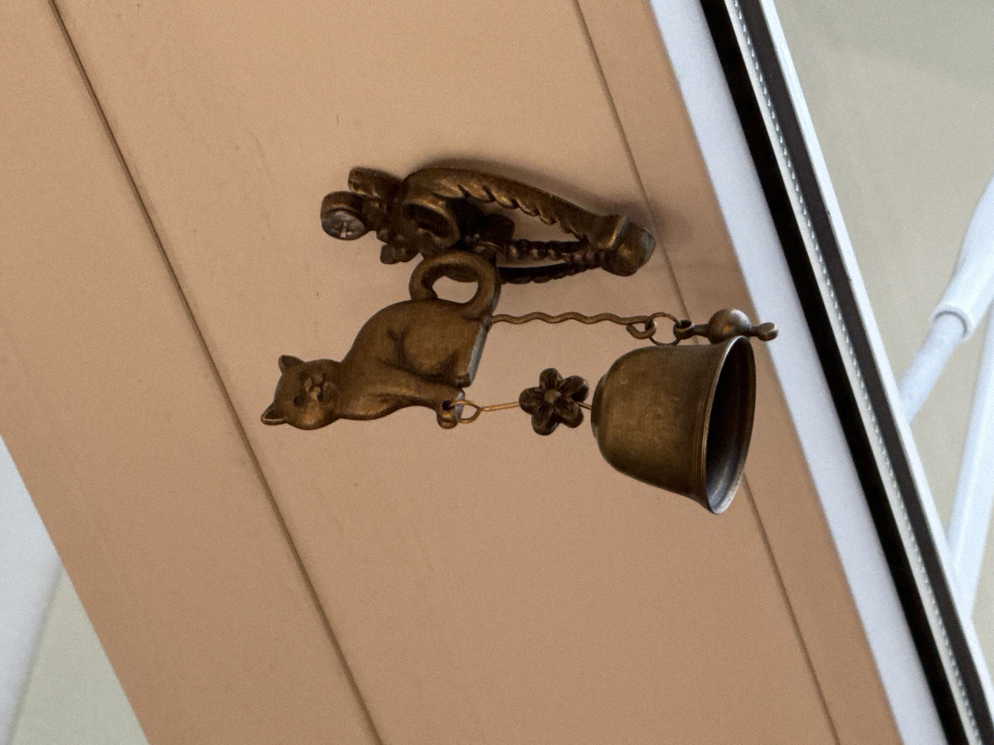 A cat-shaped door knocker with a hanging bell is mounted on a light tan, possibly wooden, surface. The knocker is made of bronze or brass and features a reclining cat figure. A floral detail is present on the chain connecting the cat to the bell. The background shows a white frame with a black stripe, suggesting the knocker is mounted near a door or window. The location is in Saint Petersburg, Russia.