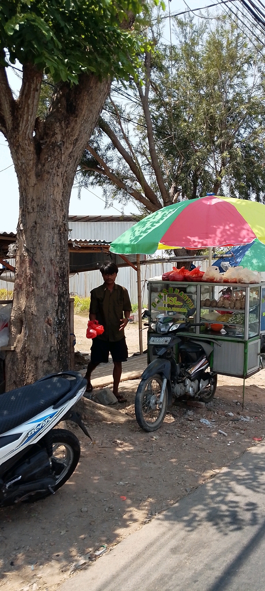 A young man with dark hair and a mustache stands in front of a food cart, holding a red plastic bag. He is wearing a dark green short-sleeved shirt and black shorts. The food cart is green and white, with a glass display case filled with food items and an umbrella overhead with red, yellow, and green panels. A motorcycle is parked to the right of the food cart, and another motorcycle is partially visible on the left. The scene is set outdoors, with a large tree trunk on the left and a fence or building in the background. The ground is a mixture of dirt, gravel, and some patchy vegetation. The overall lighting suggests it is daytime, with shadows indicating the sun is overhead. The location is identified as Sukamaju, Indonesia.