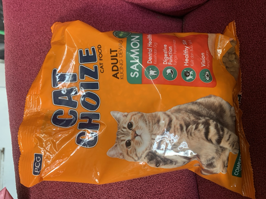 An orange bag of "CAT CHOIZE" brand adult cat food, flavored "SALMON," is displayed lying on a reddish-maroon soft surface, likely an armchair or rug, in Metro, Indonesia. The bag features a realistic image of a light brown tabby cat looking directly forward with wide eyes.
The packaging highlights several benefits for cats, written in both English and Indonesian: "Dental Health Kesehatan Gigi," "Digestive Function Fungsi Pencernaan," "Healthy Skin Kulit dan Bulu Sehat," and "Vision Penglihatan." Each benefit is accompanied by a small icon. The top right corner of the bag is slightly open, revealing dry cat kibble inside. Other visible text includes "PCG" on the bottom left, "CAT FOOD," "ADULT KUCING DEWASA," and partially visible words like "GUARANTEED" and "COMPLETE" on the right side of the bag. The bag is made of glossy plastic, reflecting some light. No people are visible, and there is no specific activity taking place, as the object is static. The setting appears to be indoors, possibly a domestic environment, with ambient lighting suggesting daytime.