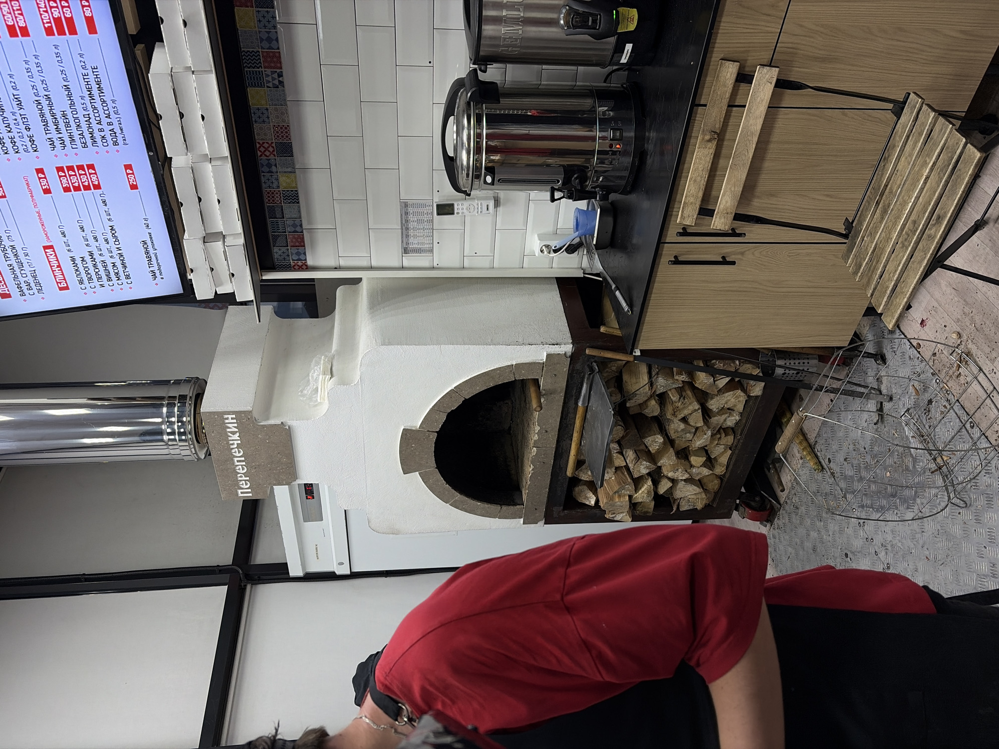 The image shows an indoor setting that appears to be a small cafe or food establishment in Izhevsk, Russia. The main subjects are a menu board with prices in Russian rubles, food preparation equipment, a wood-fired oven, and a person partially visible in the foreground.
To the left, a vertical menu board displays a list of items such as coffee, blinchiki (pancakes), and tea, with corresponding prices in Russian rubles. Adjacent to the menu board is a cabinet with food packaging.
In the center of the frame are stainless steel beverage dispensers on a black countertop. Below them is a white-painted oven with a stone archway, filled with firewood. The oven seems to be the primary feature for cooking. To the right of the oven, there is a metal rack of additional firewood. A wooden folding chair is next to the counter, and a metal grate is on the floor nearby.
A person wearing a red shirt and black pants is partially visible in the foreground, obstructing a clear view of the lower part of the scene. The floor appears to be covered with textured metal plating. The overall setting conveys a rustic yet functional atmosphere.