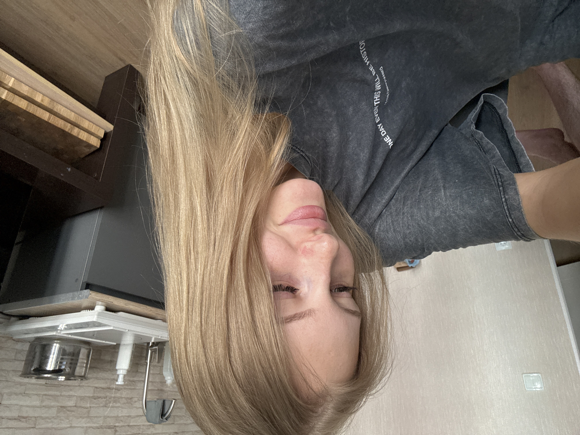 A young woman with long blonde hair is pictured indoors. She is wearing a dark grey t-shirt with text printed on it. Her head is tilted down and to the side, and she is looking down. The background shows a textured wall and a wooden floor. On a shelf in the background are several objects, including what appears to be a kettle and bottles. The lighting suggests an indoor environment, possibly a domestic setting. The location is not identifiable as Perm, Russia, based on the visual information provided.