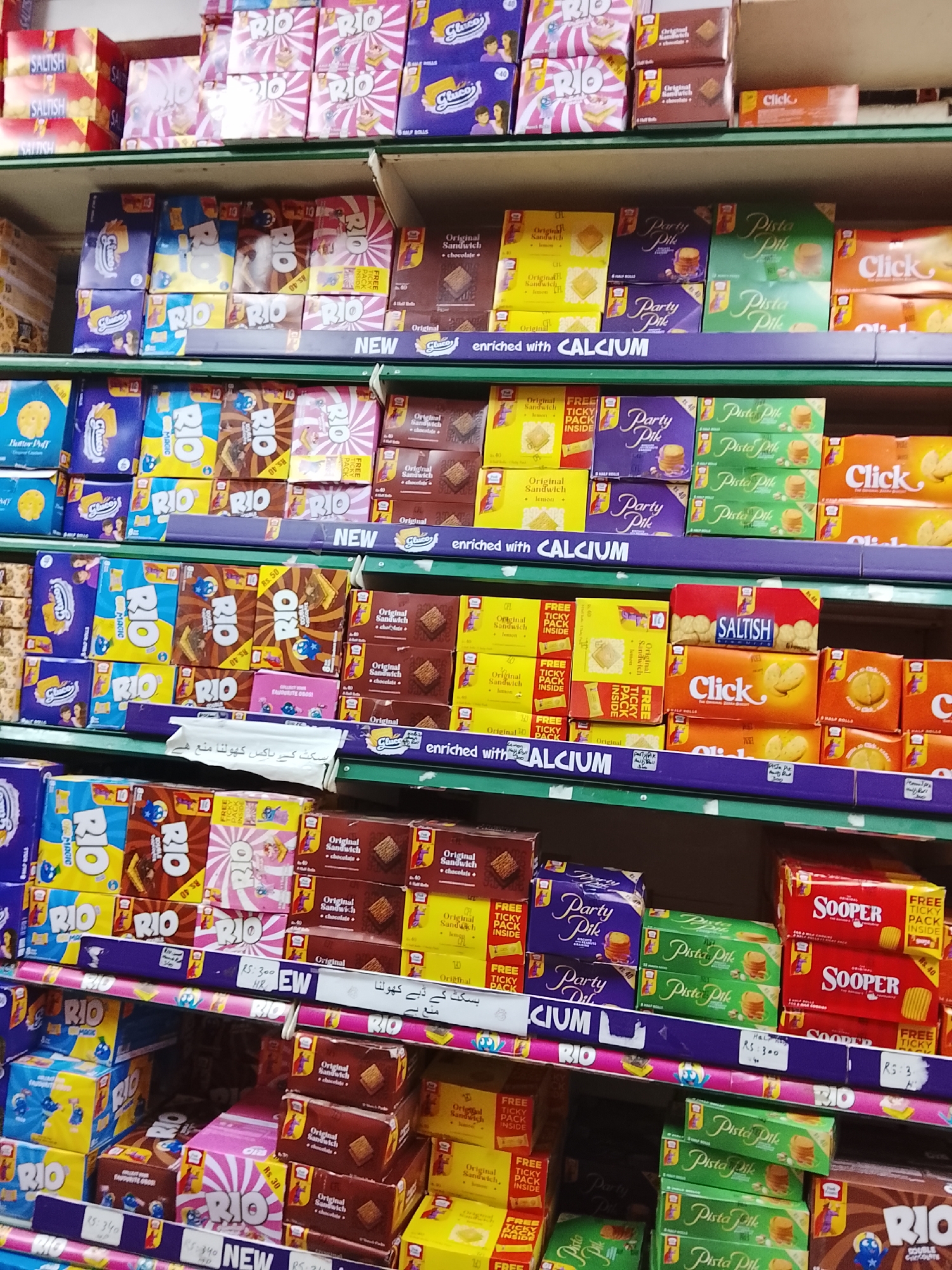 The image shows a shelf in a shop in Gulgasht Colony, Pakistan. The shelves are stocked with a variety of packaged biscuits and cookies. Brands such as RIO, Gluco, Click, Party Pile, Pista Pile, and Sooper are visible. The biscuits are arranged in stacks, with some displays labeled as "NEW" and indicating promotional offers such as "FREE TICKY PACK INSIDE". Pricing, indicated in Rupees (Rs), is visible on some of the products. A sign written in Urdu warns against opening the biscuit boxes. The shop appears to be well-stocked, and the biscuits are the main focus of the image. The image is taken from a relatively close perspective, offering a clear view of the products and their packaging. The shelves are illuminated, likely by artificial lighting.
