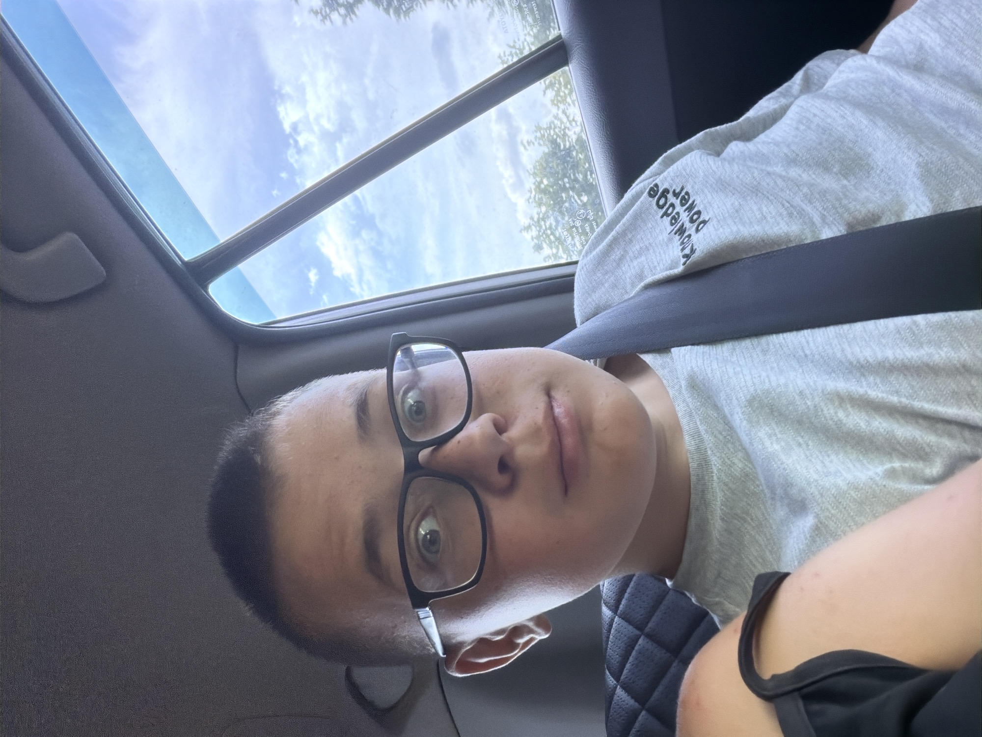 A young boy with short hair and black-rimmed glasses is sitting in the passenger seat of a car, looking directly at the camera. He is wearing a grey t-shirt with the words "Knowledge Power" printed on the sleeve. The seatbelt is visible across his chest. The background shows the interior of the car, including the car seat and the window with a view of the sky and trees outside. There is no information in the image to indicate the location is Kruglovka, Russia.