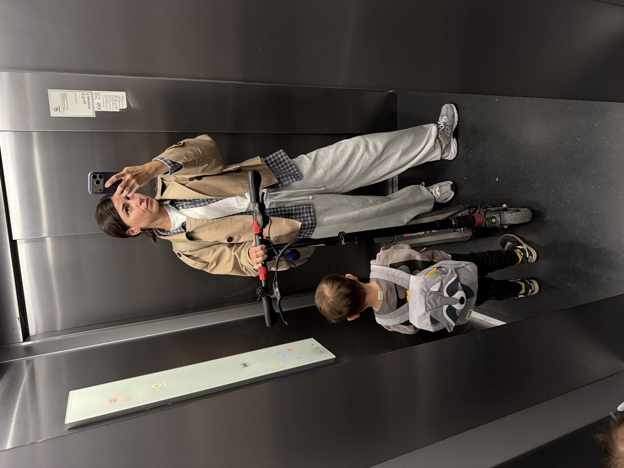 The photo shows a woman and child in an elevator. The woman is taking a selfie with her phone while holding the handlebars of a folded scooter. She is wearing a trench coat, plaid shirt, grey sweatpants, and New Balance sneakers. The child is wearing a raccoon backpack and black pants with yellow accents. They are standing on a black floor in an elevator with stainless steel walls and a panel of buttons. There is a notice posted on the wall near the woman's head. This photo was taken inside a shopping center in Budva, Montenegro.