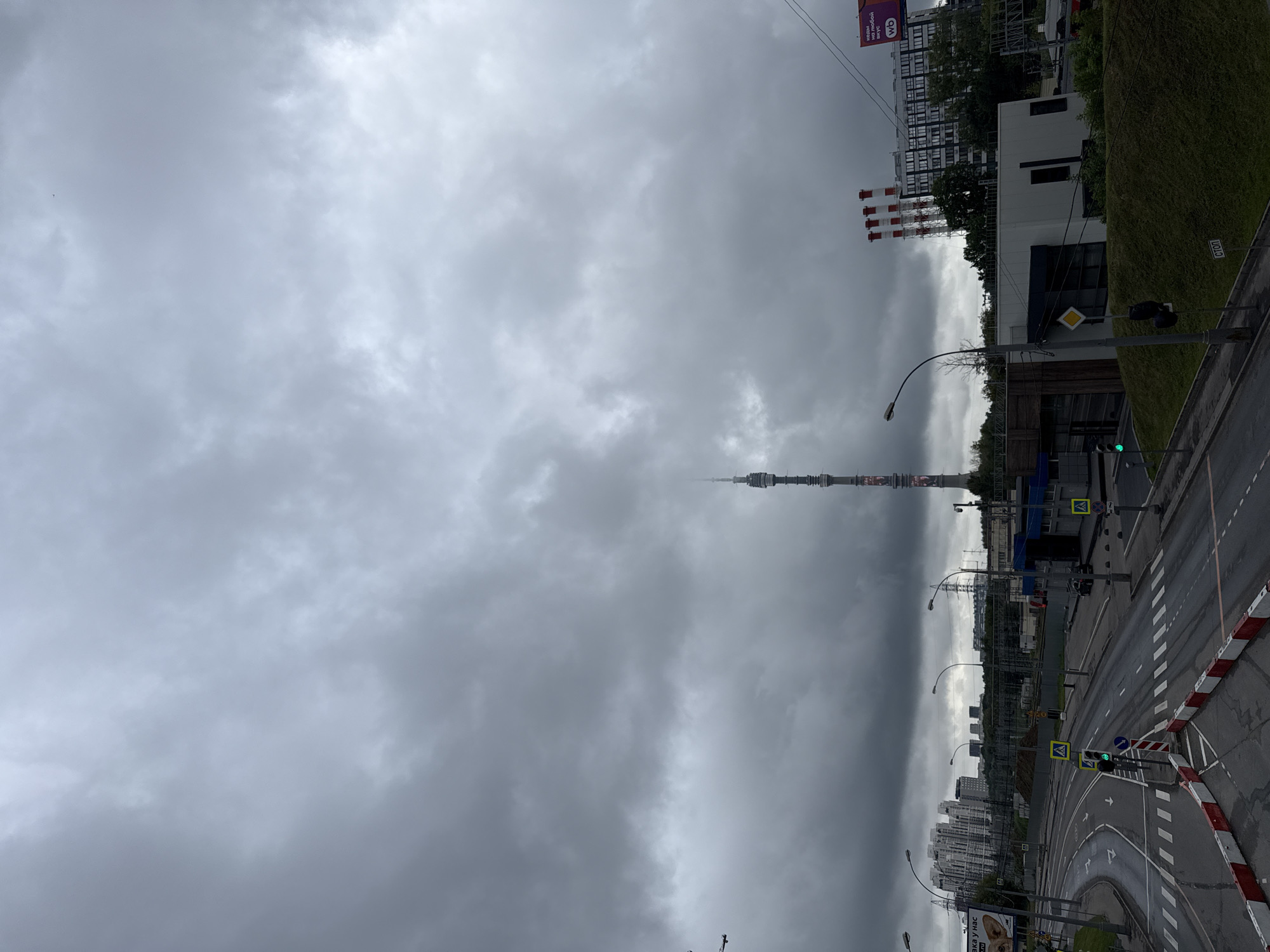 The image captures a view of the Ostankino Tower in Moscow, Russia, under a cloudy, overcast sky. In the foreground, a road intersection with pedestrian crossings is visible. Traffic lights, road markings, and barriers are present. To the left, apartment buildings and infrastructure can be seen. In the midground, there are industrial buildings, including a power plant with red and white smokestacks. The Ostankino Tower, a prominent landmark, stands tall in the distance. The overall scene depicts an urban environment with functional infrastructure and towering structures, set against a somber sky.