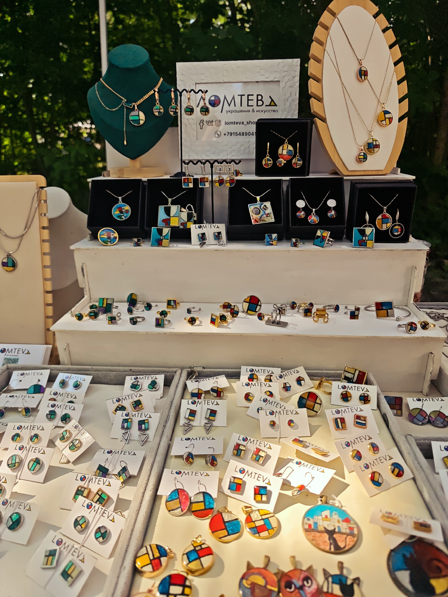 This image shows a display of artisanal jewelry at an outdoor market. The jewelry, primarily necklaces and earrings, features colorful geometric designs reminiscent of Piet Mondrian's work. The items are displayed on tiered white stands and within black gift boxes. Some pieces are presented on white cardboard cards with a logo that reads "LOMTEVA" in Cyrillic. In the background, there is a green velvet mannequin displaying a necklace and earrings, and a wooden display stand with several necklaces. The overall setting appears to be a sunny day, with trees and foliage visible in the background. The presence of the logo and the style of the jewelry suggest this is likely a vendor's booth at a craft fair or market. Based on the Cyrillic text and the style of the jewelry, this market is likely located in Russia. The specific location in the city of Kimry, Russia cannot be determined from the image alone, but it is plausible given the context of such markets in Russia. There are no people visibly present in the image, but the setup implies a commercial transaction is taking place or about to take place.