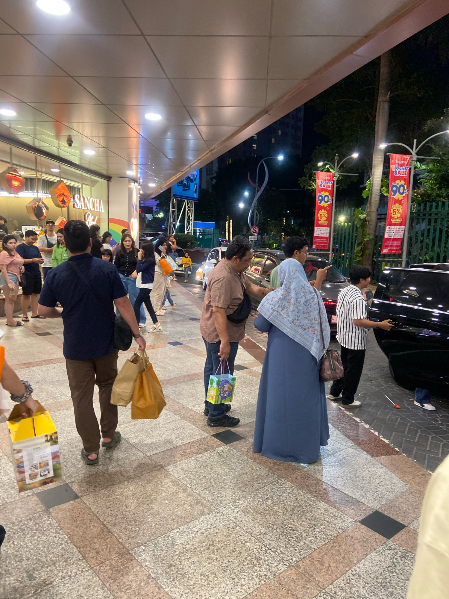 This image depicts a nighttime scene outside a commercial establishment in Surabaya, Indonesia, possibly a shopping mall or a retail store. The exterior features a covered walkway with recessed lighting. Two prominent red banners with white text advertising a sale of "90%" and "5%" are visible, indicating a commercial event. Several individuals are present, some carrying shopping bags. In the foreground, a man in a dark blue shirt and brown pants is seen from behind, carrying two large brown paper shopping bags. To his right, a woman in a blue hijab and dress is interacting with a person whose arm is partially visible. A young boy in a striped shirt stands near a black car. Other shoppers are scattered throughout the scene, some walking and others standing. The ground is paved with patterned tiles. The presence of cars and the general ambiance suggest an urban commercial setting.