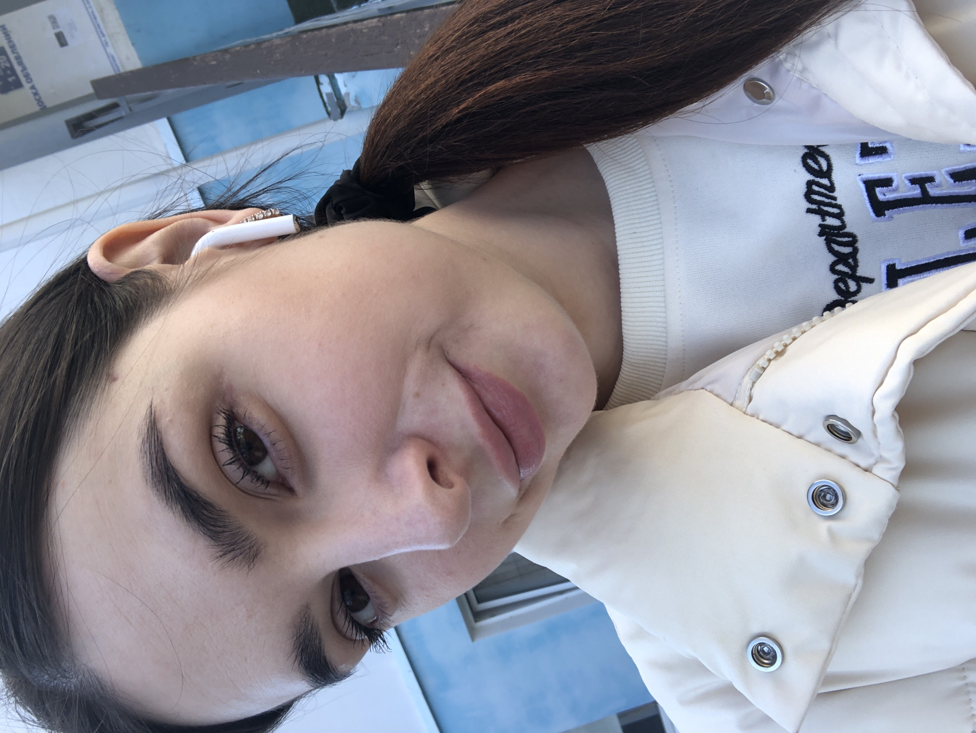 A young woman poses for a selfie outdoors. She is wearing an off-white sweater with "department" embroidered on it, covered by an unzipped off-white puffer jacket. Her dark brown hair is pulled back in a ponytail with a black hair tie. An AirPod-style wireless earbud is visible in her left ear. The background shows a blue sky and the exterior of a building. The location may be Chaikovsky, Russia.