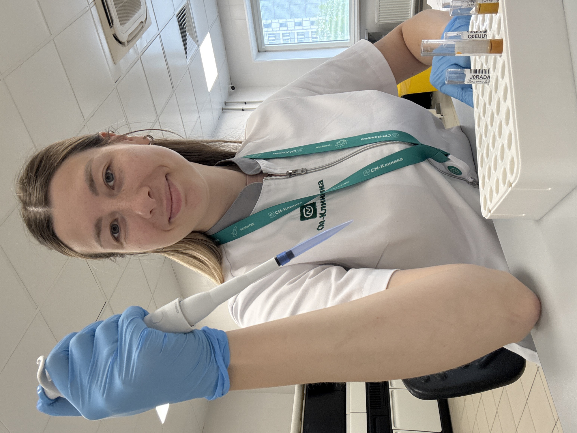 A Caucasian woman with blonde hair, wearing a white lab coat and blue nitrile gloves, is seated at a table in a laboratory. She is holding a pipette in her gloved right hand, poised over a rack of test tubes containing a brown liquid. Her left arm is bent, resting on the table. The background shows laboratory equipment, including a large machine on the left and windows with a view of a city, likely Moscow, Russia, on the right. The woman is looking towards the camera with a slight smile. The lighting suggests an indoor, well-lit environment.