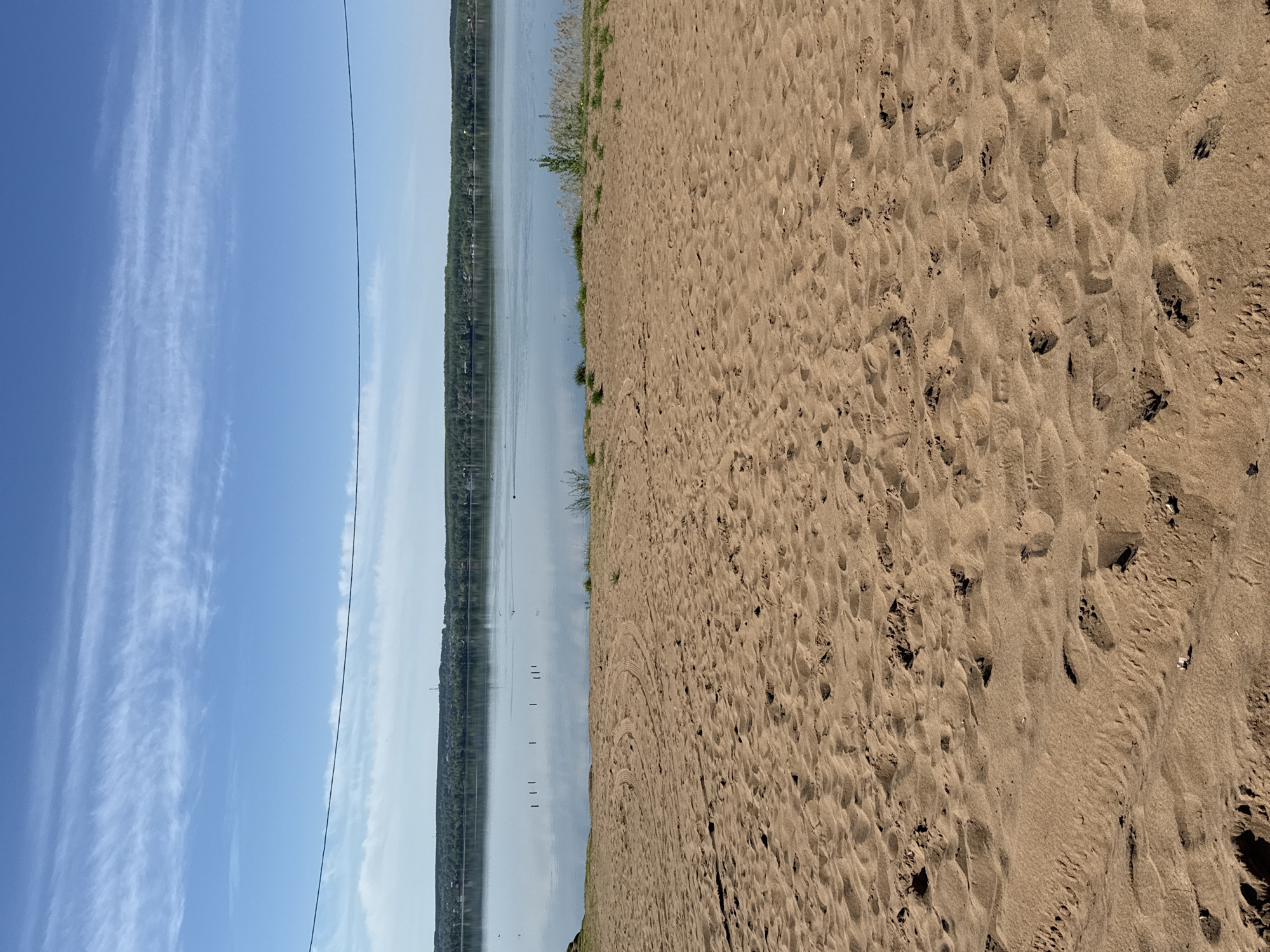 The image shows a beach scene in Votkinsk, Russia. On the right, there is a sandy beach with multiple footprints and disturbed sand patterns. The sand is dry and light brown in color. To the left of the beach, there's a body of water, likely a lake or a wide river, reflecting the sky and distant shoreline. The shoreline is lined with trees and some visible buildings. The sky is light blue with long, thin clouds stretched horizontally. A thin wire is visible running horizontally across the sky and water, likely a power line.