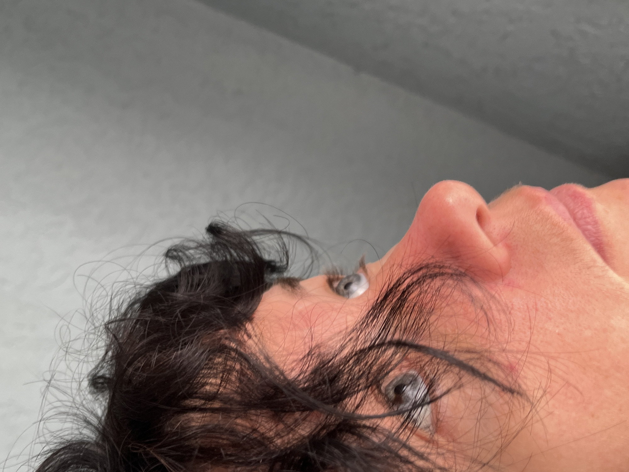 The image is a close-up, profile view of a person's face, focusing on their left eye, nose, and mouth, with a portion of dark, curly hair visible. The person appears to be looking upwards and to the left, away from the camera. The skin on their face is fair and shows some redness, particularly around the nose. The background is a plain, textured gray surface, which appears to be a wall or a similar indoor element. There is no indication of a specific location within ღრმაღელის თემი, Georgia, or any contextual environmental clues to suggest a city setting. The image is a neutral, unembellished portrayal of a facial close-up against a simple backdrop.