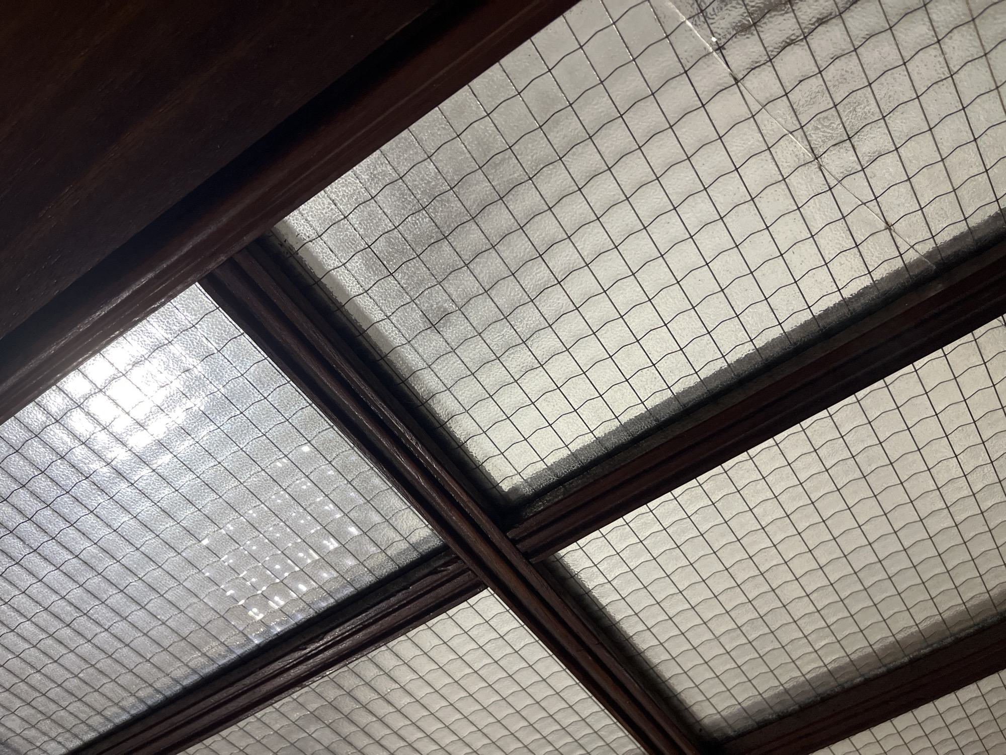 The image shows a close-up view of a window with a grid pattern. The window is framed by dark wooden beams. The glass is textured and reflects light, creating a shimmering effect. There are no people or animals visible in the image. The scene appears to be indoors, possibly a home or a commercial establishment, given the architectural elements. The lighting suggests it's daytime, with sunlight filtering through the window. The overall impression is one of textured glass and dark wood, conveying a sense of depth and subtle detail. The location of Juan Griego, Venezuela, is mentioned but not depicted in the image itself.