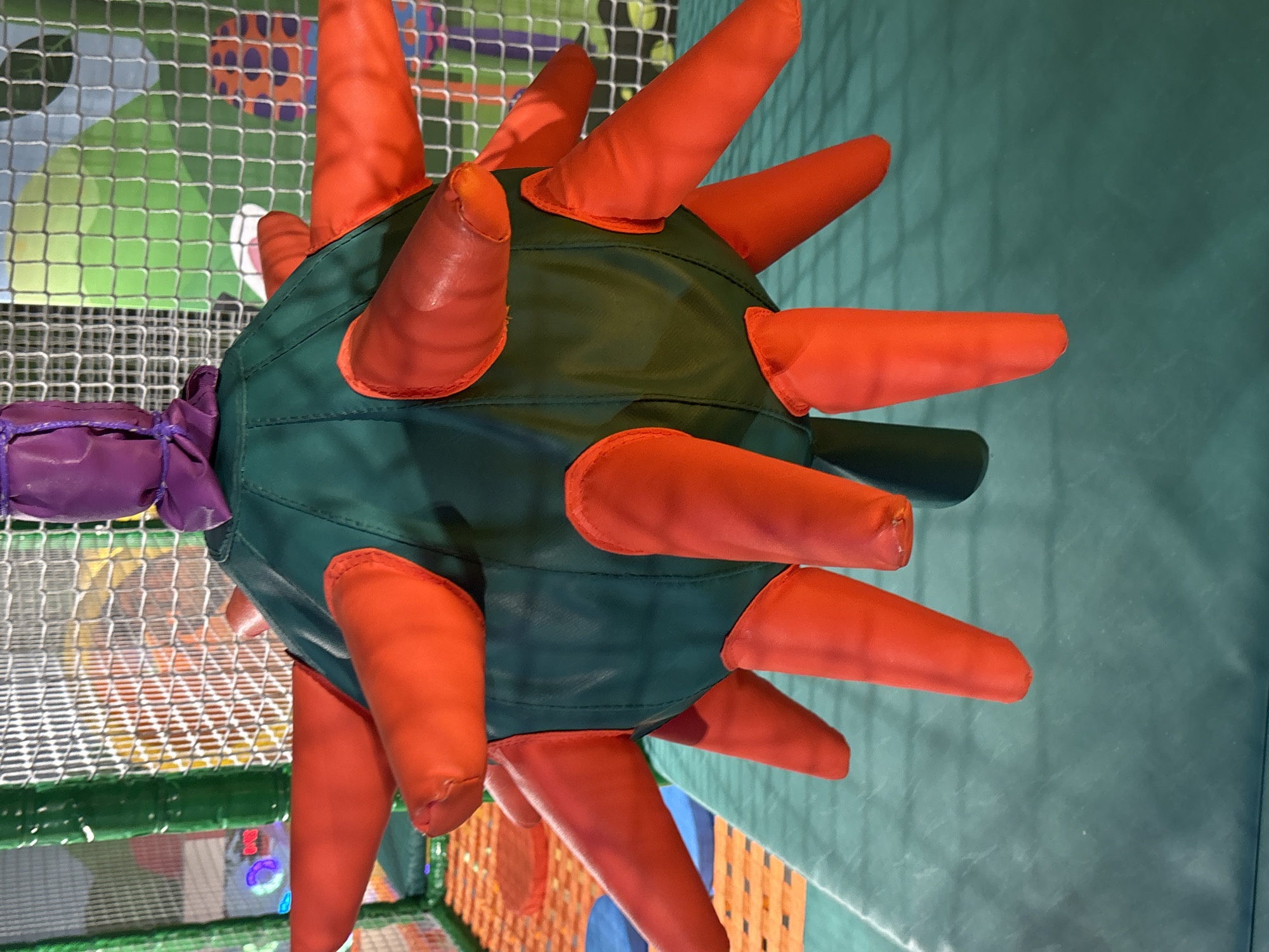 A close-up, head-on shot captures a plush, ball-shaped toy with numerous red, finger-like appendages protruding from its dark green surface. The toy is suspended from a purple fabric knot, adding a splash of color. The background features a white mesh net, suggesting an indoor play area. The floor is a solid, dark green mat, and beyond the net, a colorful, soft play structure with orange and blue elements is visible. The lighting appears to be natural or indoor ambient light, creating soft shadows and highlighting the texture of the toy. The setting is likely an indoor children's play center in Dubai, United Arab Emirates, a common type of recreational facility in the city.