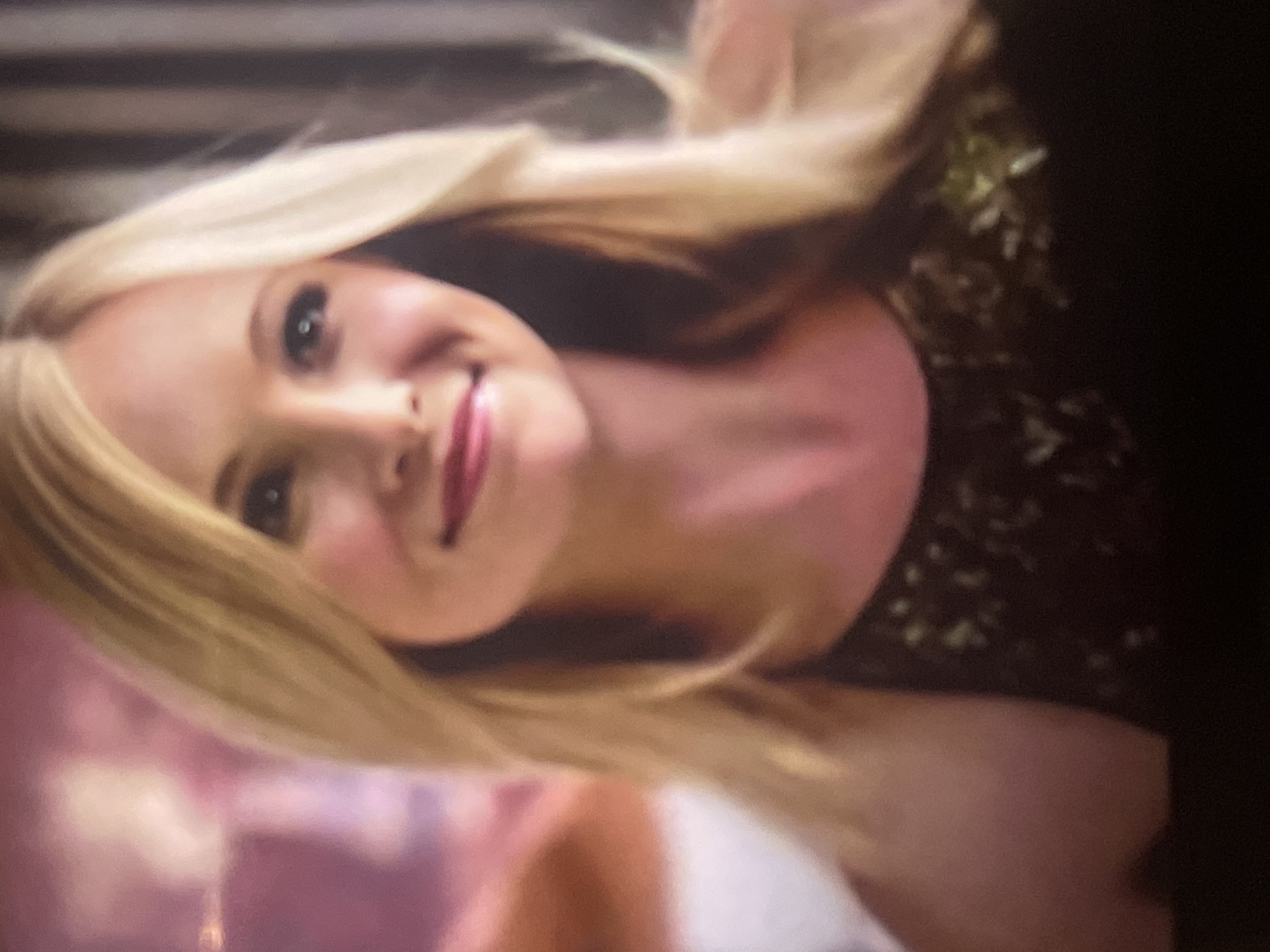 This image features a blonde-haired woman with fair skin and light-colored eyes, positioned at a tilt. She is looking directly at the viewer with a slight smile or smirk, conveying a happy and playful emotion. Her cheeks appear flushed. She wears a dark-colored top, possibly a dress or shirt, adorned with subtle, lighter-colored, potentially sparkly or embroidered, details on the upper left side of her torso.
The background is largely out of focus. On the left side, there's a soft blur of pink and purple hues, while the upper left shows darker, possibly striped, elements. The lighting suggests an indoor setting, likely under artificial light. There are no clear location cues within Poltava, Ukraine, visible in the blurred background, nor any discernible weather conditions. The woman is the main subject, seemingly posing for a photograph. No text is visible in the media file.