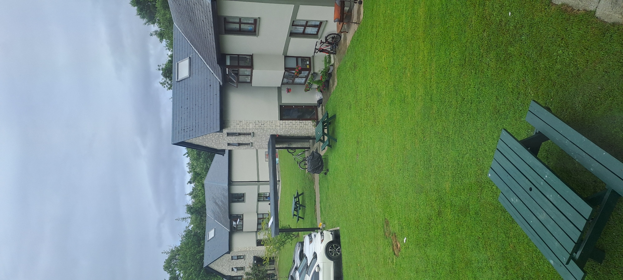 The image shows a view of a residential area in Castlebar, Ireland, on an overcast day. In the foreground, a green picnic table is visible on a manicured lawn. Beyond the lawn, there are several apartment-style buildings with light-colored facades and dark-tiled roofs. Cars are parked in a lot to the left of the buildings. In the yard area between the buildings, there are more picnic tables, a charcoal grill, and several bicycles. The buildings appear to be two or three stories tall and are surrounded by trees and greenery. The overall scene depicts a quiet, suburban residential setting.