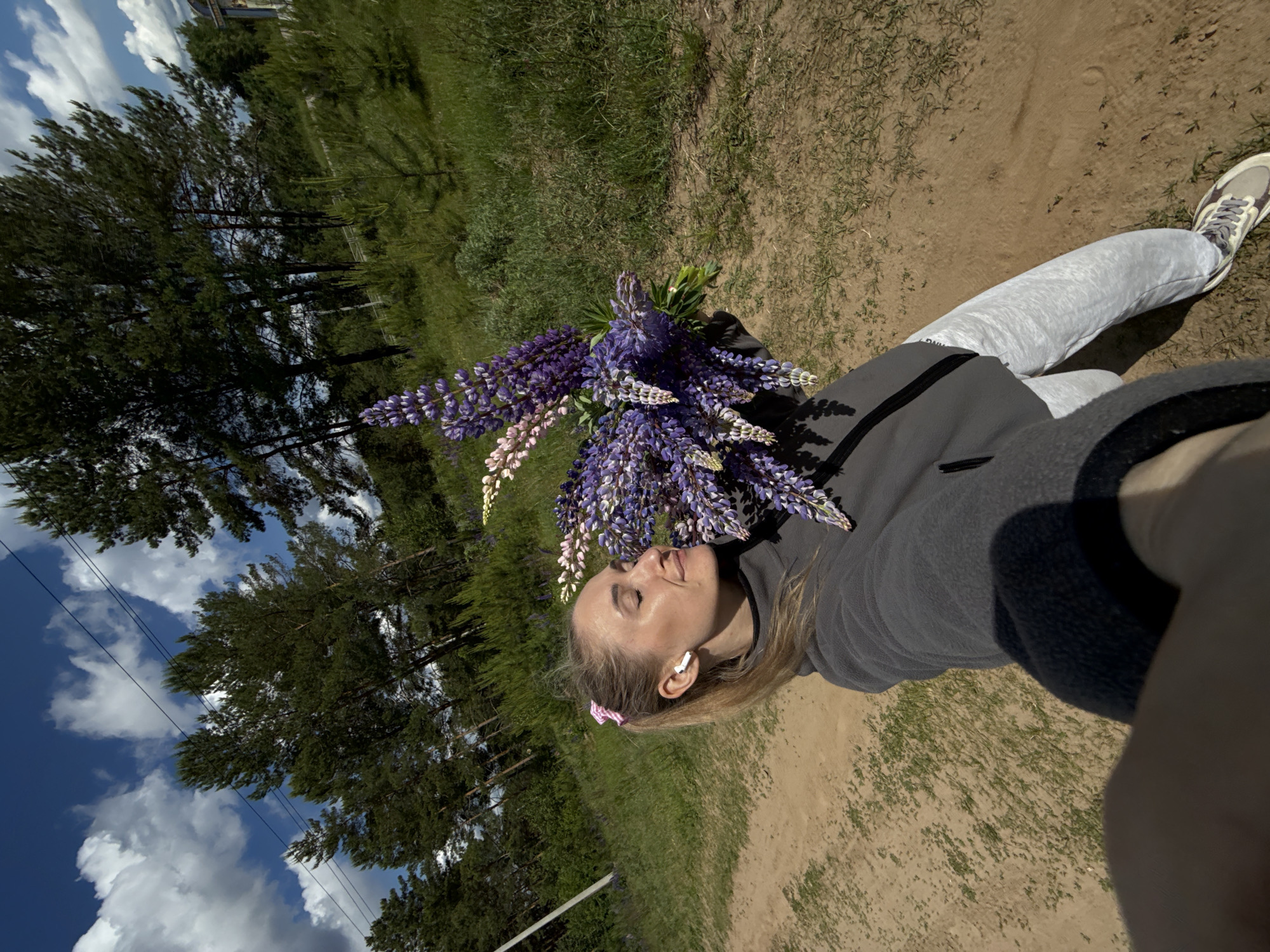 The image shows a woman lying on a dirt and grassy area near trees, possibly a park or roadside in a rural setting. She is wearing light gray sweatpants, a gray sweatshirt, and light-colored sneakers. She has blonde hair, a pink hair accessory, and is wearing an earpiece. She is holding a large bouquet of purple and white lupine flowers. The photograph appears to be a selfie, with part of the photographer's arm visible in the corner. The background includes trees, grass, a blue sky with puffy white clouds, and what might be power lines. The location is likely Chaikovsky, Russia based on the prompt's instructions.