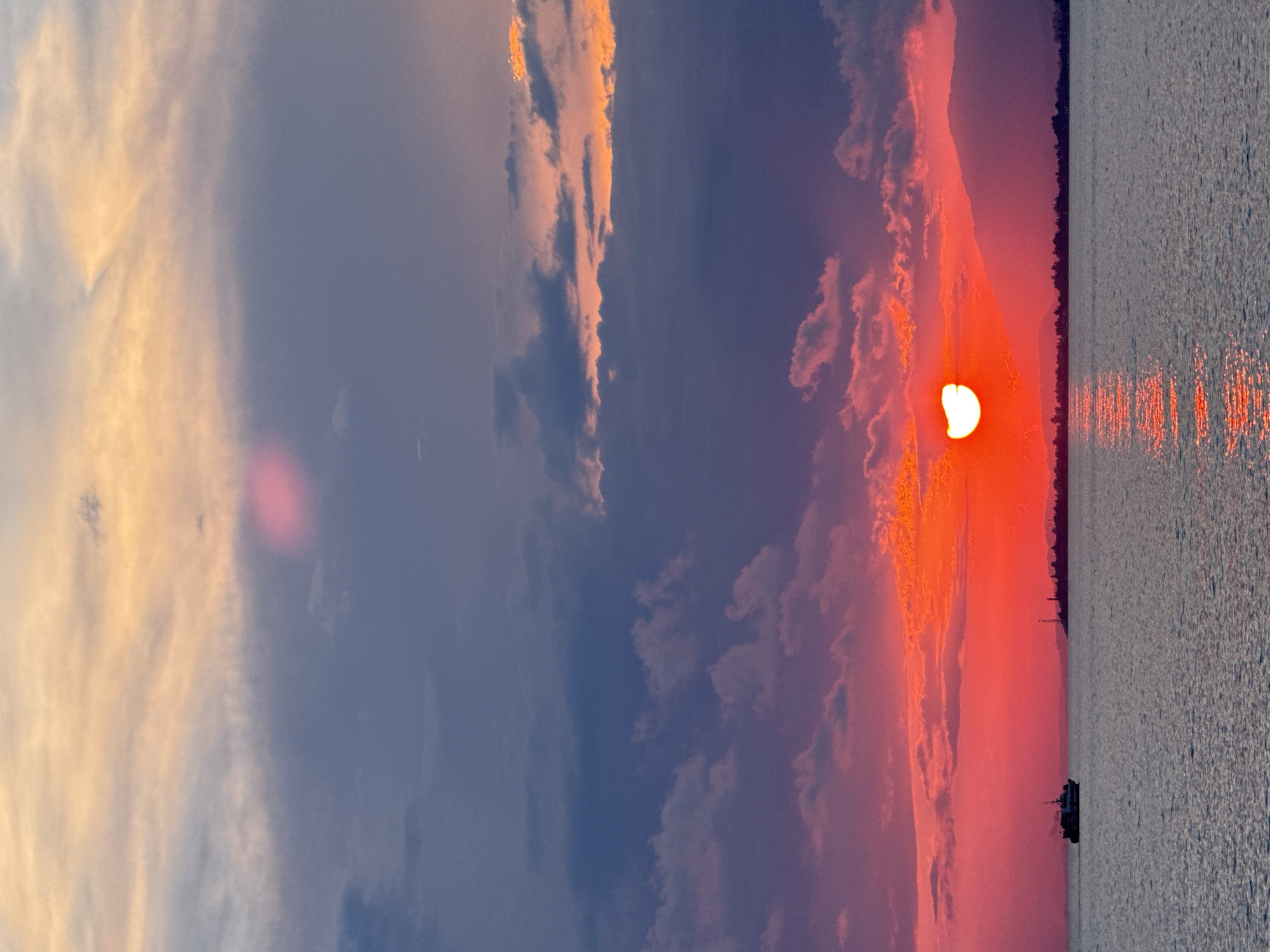 An intense sunset paints the sky over the sea in the city of Yuada-Gulrypsh, Abkhazia. The sun, a vibrant red orb, is partially obscured by clouds, casting a fiery glow across the horizon. The sky transitions from brilliant orange and pink near the sun to deep purple and blue higher up, with scattered clouds adding texture and depth. In the foreground, the calm sea reflects the colors of the sunset, creating a shimmering path of light. A small boat sails peacefully on the water, silhouetted against the dramatic sky. A distant shoreline is visible, marked by a faint silhouette of mountains and a small lighthouse. The overall atmosphere is one of serene beauty and dramatic natural spectacle.