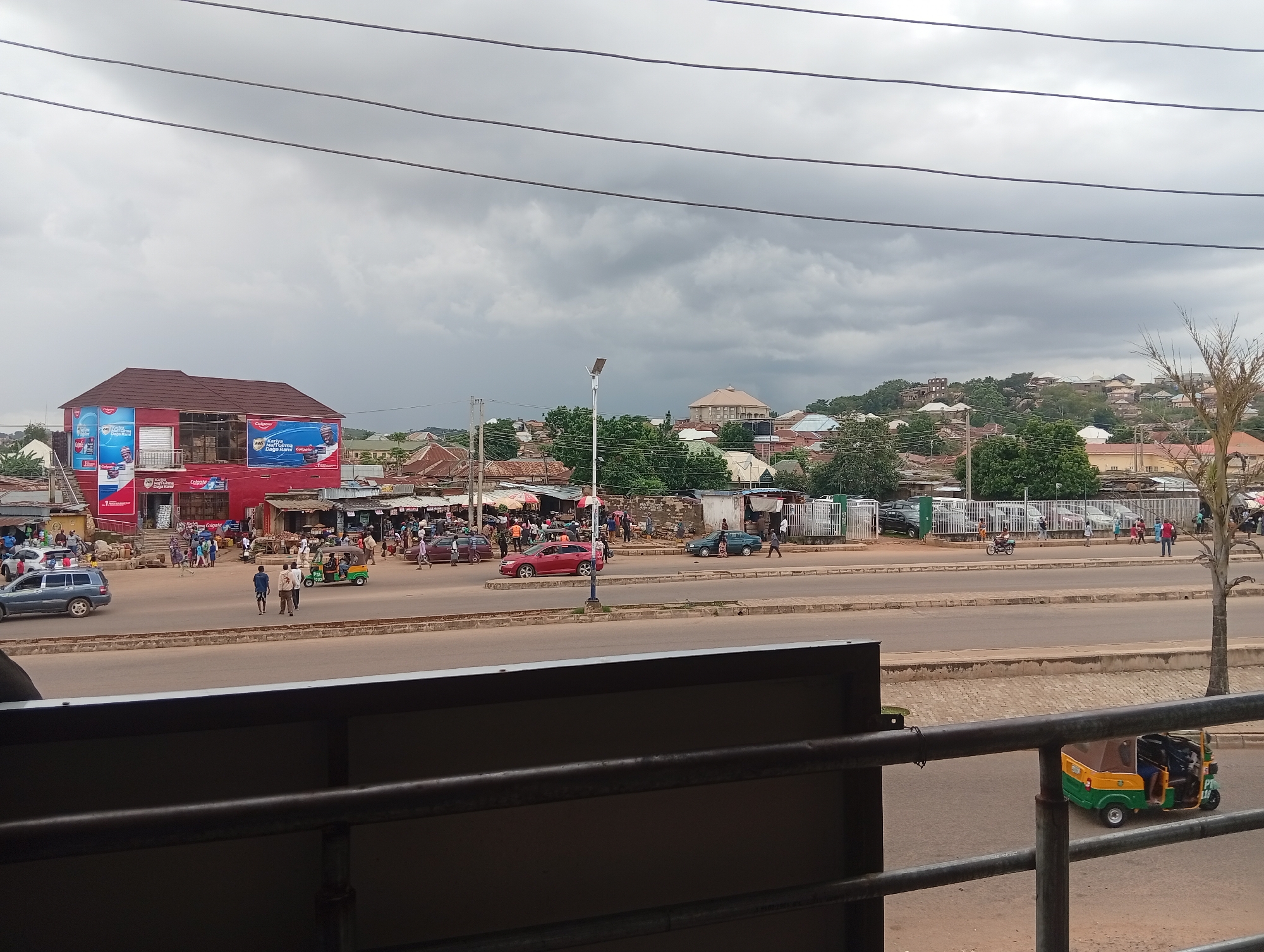 The image shows a street scene in Kabong, Nigeria. The foreground features a dark metal railing, suggesting the photo was taken from a balcony or elevated vantage point. Beyond the railing is a paved road with a marked lane separation.
On the right side of the road, a green and yellow auto-rickshaw, commonly known as a "Keke Napep," is visible, moving away from the camera.
Across the road, on the left side, there is a large red building with a white awning and large advertisements. The building appears to be a commercial establishment. In front of this building, there are several people walking and gathered, and various vehicles are parked or moving, including a dark blue SUV on the far left. Further down the street, there are more buildings, market stalls with awnings, and a mix of cars and people.
The background reveals a residential area with numerous houses, many with corrugated metal roofs, situated on a hilly terrain. The sky is overcast with heavy grey clouds. A tall, thin street lamp is visible in the middle ground, and utility wires are strung across the upper portion of the image. The overall impression is that of a bustling urban environment under a cloudy sky.
