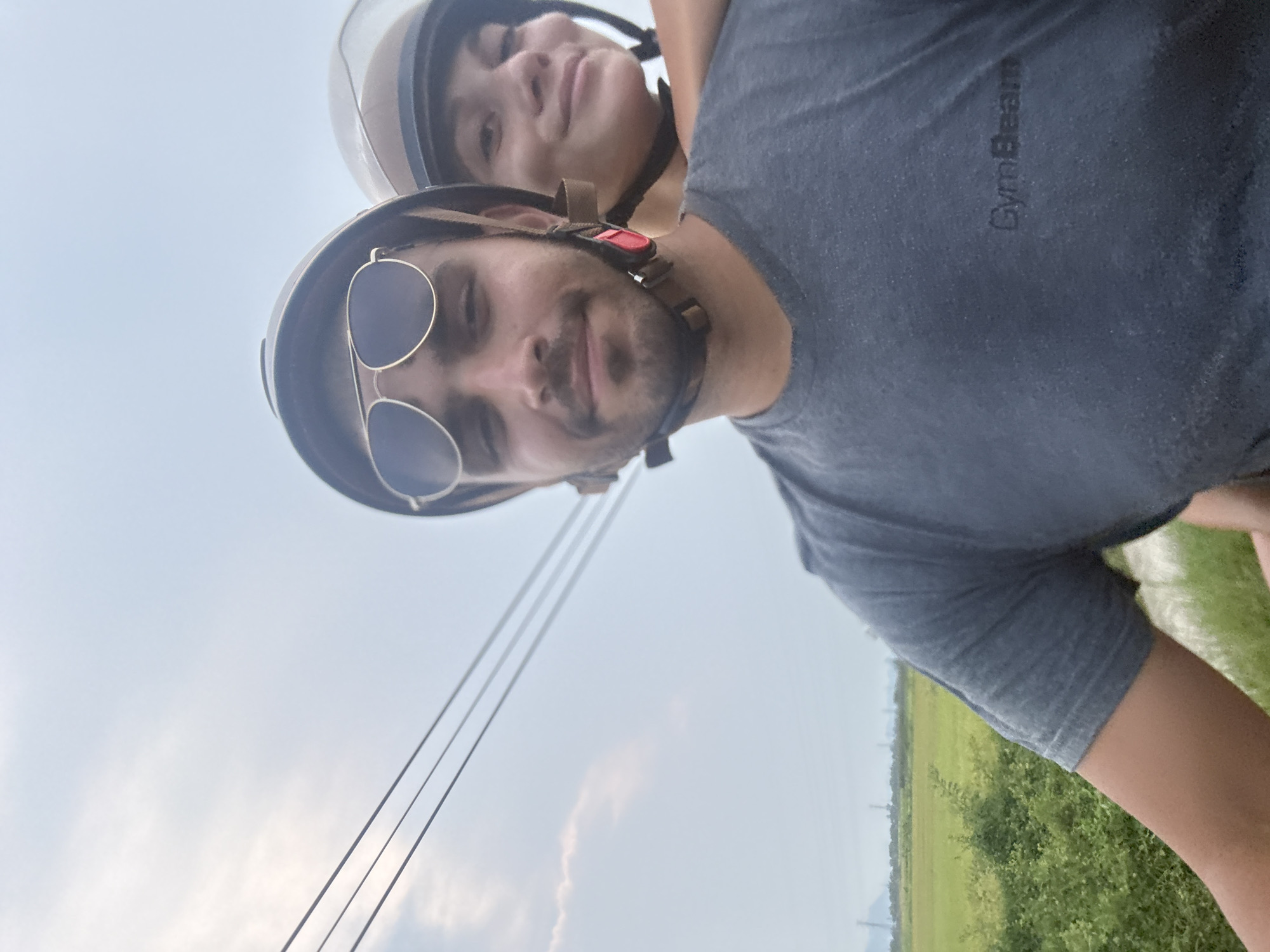 The image captures a selfie-style shot of two individuals, a man and a woman, riding a scooter or motorcycle in Đà Nẵng, Vietnam, during daytime. The perspective suggests the photo was taken from a helmet-mounted camera or held low by one of the riders.
In the foreground, a man with a beard and mustache is prominently featured, looking directly at the camera with a gentle smile. He is wearing aviator-style sunglasses and a dark-colored open-face helmet with a visible chin strap. He sports a dark grey t-shirt, which has the word "GymBeam" subtly visible on the chest. Behind him and slightly to his right, a woman is partially visible, also wearing an open-face helmet and looking towards the camera with a faint smile.
The setting is an open, rural or semi-rural landscape. The background features a wide expanse of light blue to hazy sky with scattered, soft clouds. Below the horizon, lush green fields or dense vegetation stretch out, suggesting a scenic countryside. Power lines are visible diagonally across the upper left portion of the sky, adding a touch of infrastructure to the natural backdrop. The weather appears bright but possibly slightly overcast or hazy. The overall impression is one of a leisurely ride, capturing a pleasant moment outdoors.