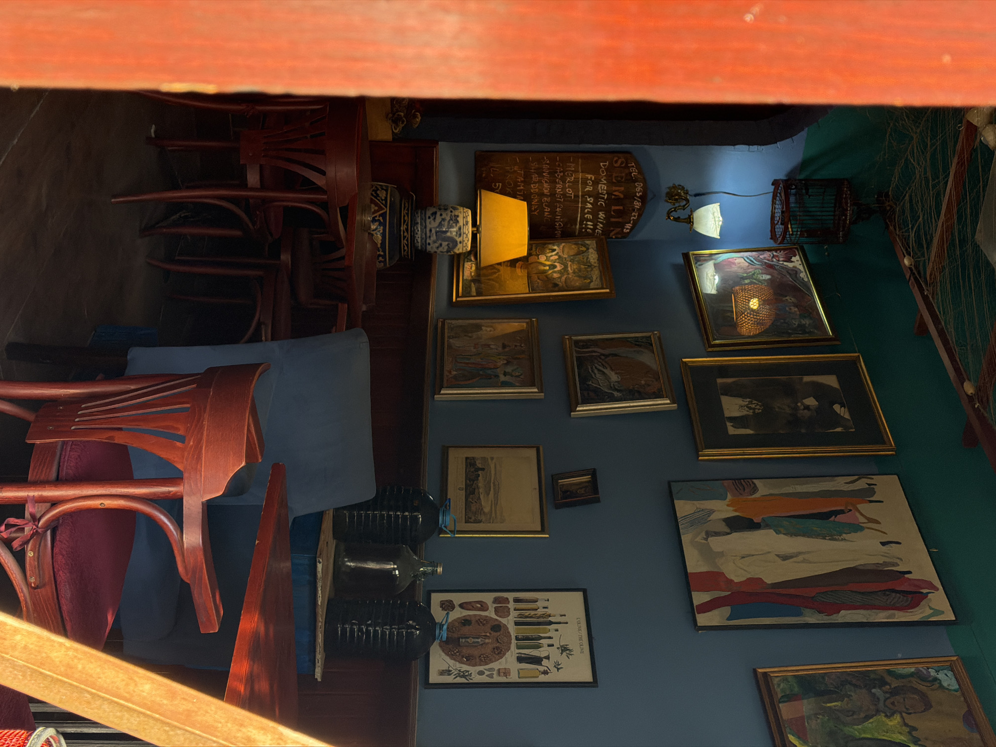 The image captures the interior of a business, potentially a restaurant or wine shop, located in the Palilula Urban Municipality, Serbia. The space is decorated with numerous framed pictures hanging on a blue wall. Some of the pictures appear to be art, while one contains information about "Semlin Domestic Wine for Sale," listing types such as Merlot and Chardonnay. Another picture is titled "L'Olive/The Olive," suggesting a Mediterranean theme.
There are dark glass bottles, likely wine or liquor, arranged on a shelf. Wooden chairs are stacked or positioned in the scene, suggesting a dining area. A table lamp with a yellow lampshade illuminates part of the wall. Additional decorative items such as a birdcage-like object and wall sconce are visible. The overall atmosphere seems cluttered yet artistic.