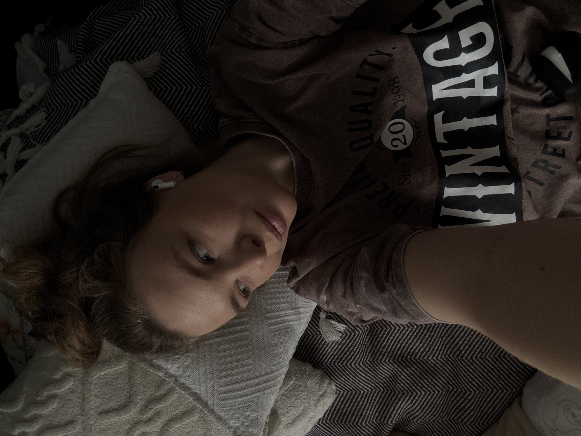 A woman lies on a bed or sofa. She wears a faded brown t-shirt with the word "VINTAGE" printed vertically in large white letters on the front. The shirt also has other smaller text and a circular design featuring the number 20 and the year 1998. A white earbud is visible in her left ear. She rests her head on a white pillow or cushion that has a textured surface. The background consists of a brown and white striped blanket and a white blanket with fringe. The woman's face is turned toward the camera. The photograph appears to be taken at close range. There is no evidence to confirm the location is Vladimir, Russia.