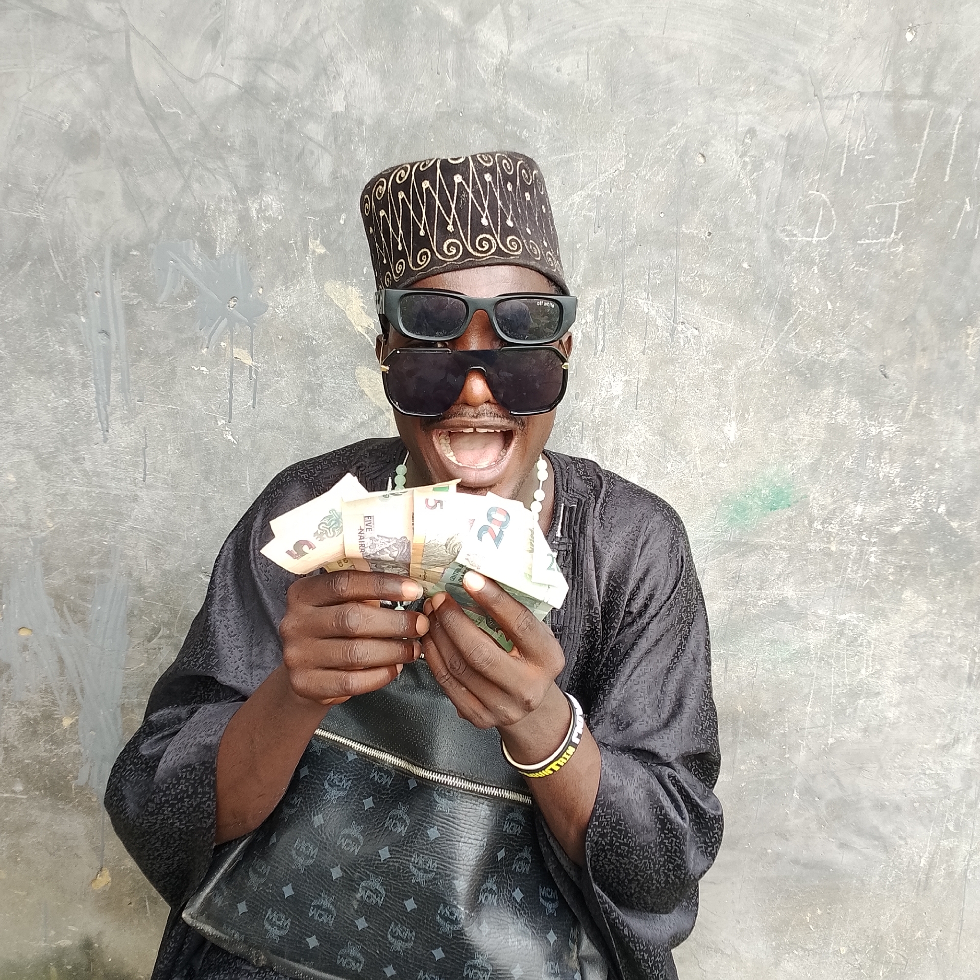 A Black male, appearing to be in his 20s or 30s, stands against a textured, light gray concrete wall. He wears a black, patterned garment and a black cap with gold embroidery. Large, dark sunglasses are worn over his eyes, and a beaded necklace is visible around his neck. He is holding a fan of Nigerian Naira banknotes in his hands and is looking towards the camera with his mouth open. A black MCM-branded bag is slung over his shoulder, and he wears a bracelet on his right wrist. The concrete wall has visible scuff marks, scratches, and some graffiti. The lighting suggests an outdoor, daytime setting.