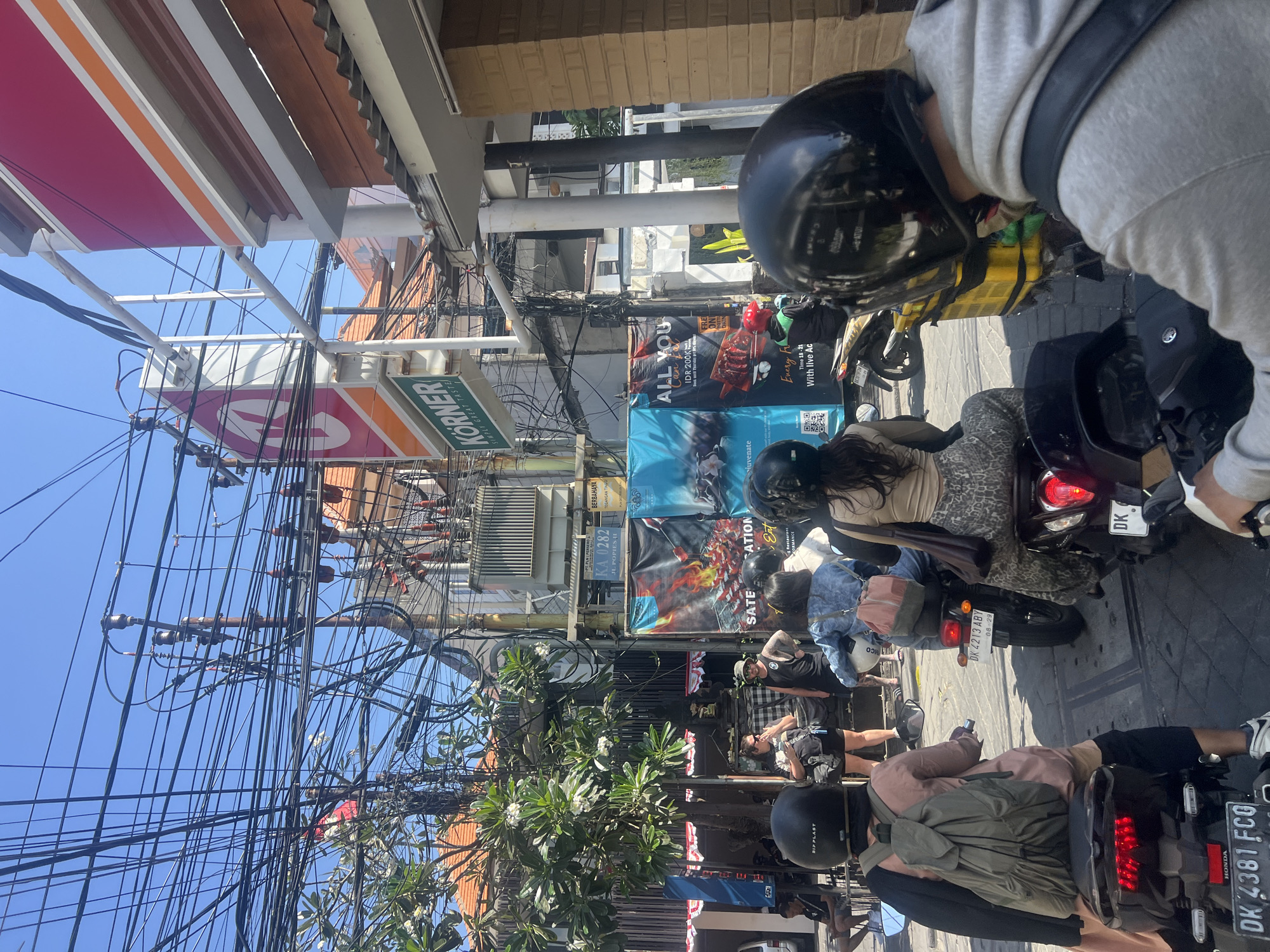 This image depicts a street scene in Kuta, Indonesia, characterized by heavy overhead electrical wiring. In the foreground, several people are seen riding motorcycles, their backs to the camera. One individual in the center wears a pink top and a green backpack, while another to their right has on a patterned skirt and a brown purse. Behind them, another person wears a denim jacket and a light pink backpack. A person on the far right is wearing a grey hoodie.
Further back, more motorcycles and people are visible, interspersed with street vendors' stalls and buildings. Notably, a sign for "KÖRNER" with a prominent red circular logo is visible on a building. Another sign displays "ALL YOU CAN EAT" with prices and details. Large banners with images of grilled food and text like "SATEFICATION" and "Enjoy Every Bite With Live Ac" are also present. A large electrical transformer is mounted on a pole amongst the dense network of wires. The street is paved, and the weather appears to be sunny with a clear blue sky. The overall impression is one of a bustling, busy urban environment.