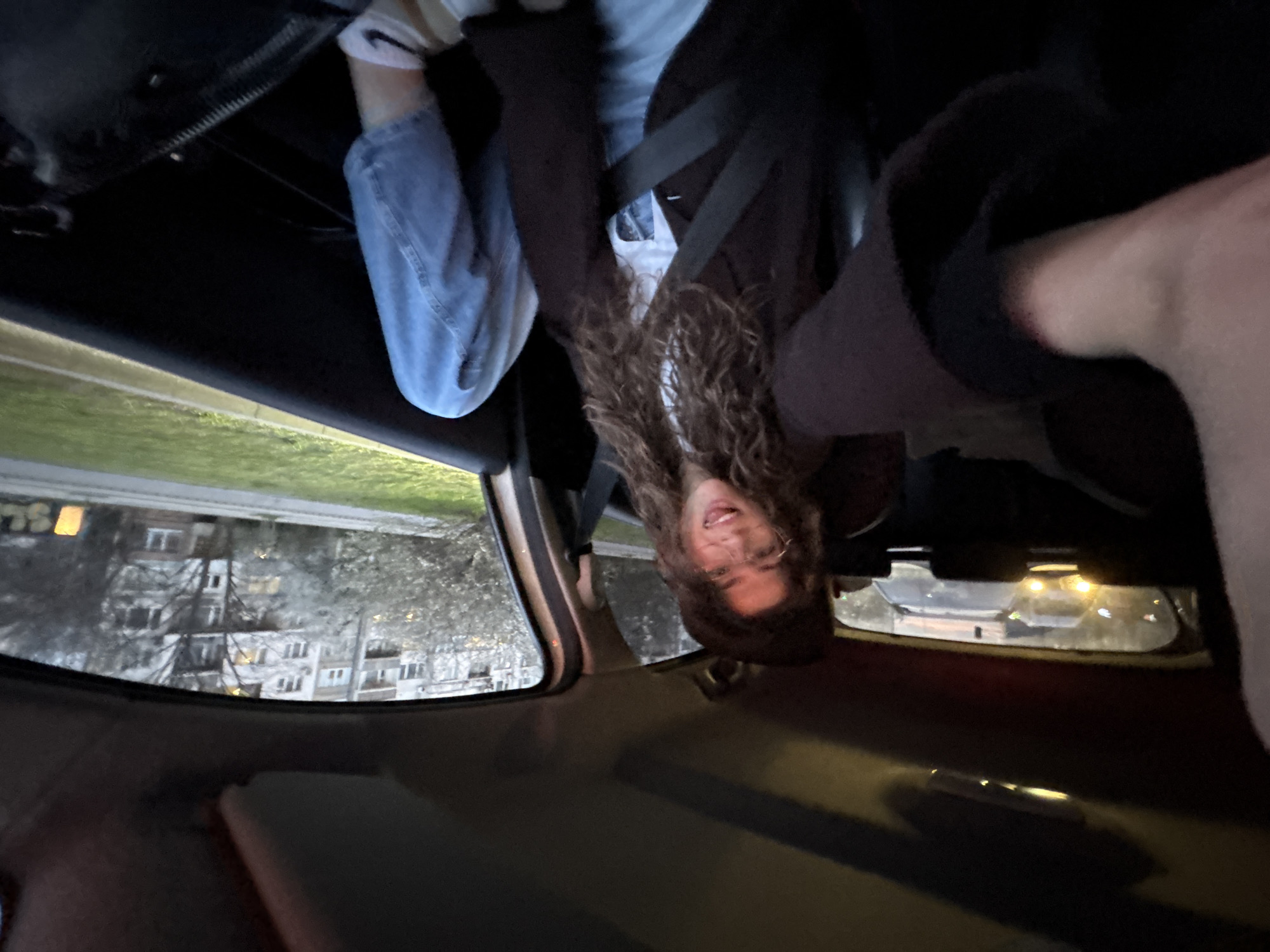 The image displays the interior of a passenger vehicle from a first-person perspective, capturing a young adult female seated in the front passenger seat and the exterior environment through the windows. The female has long, curly brown hair and wears clear-framed eyeglasses. She is smiling with an open mouth, showing her tongue slightly. She is dressed in a dark brown jacket over a white top, light blue jeans, and white socks with a black Nike swoosh logo on her right foot. A seatbelt is fastened across her chest. Her left arm is extended towards the camera, indicating she is taking a selfie.
Through the front passenger window, a paved road is visible, flanked by a green grass verge and multiple large, multi-story residential block buildings typical of socialist-era architecture in Sofia, Bulgaria. Trees, some appearing bare or sparsely leafed, are situated between the road and the buildings. Artificial yellow light sources are visible within the buildings and on the far right of the exterior scene. Through the rear window, the illuminated headlights of at least two vehicles behind are visible, suggesting the car is in traffic. The overall lighting indicates dusk or early evening. The location is an urban residential district in Sofia, Bulgaria.