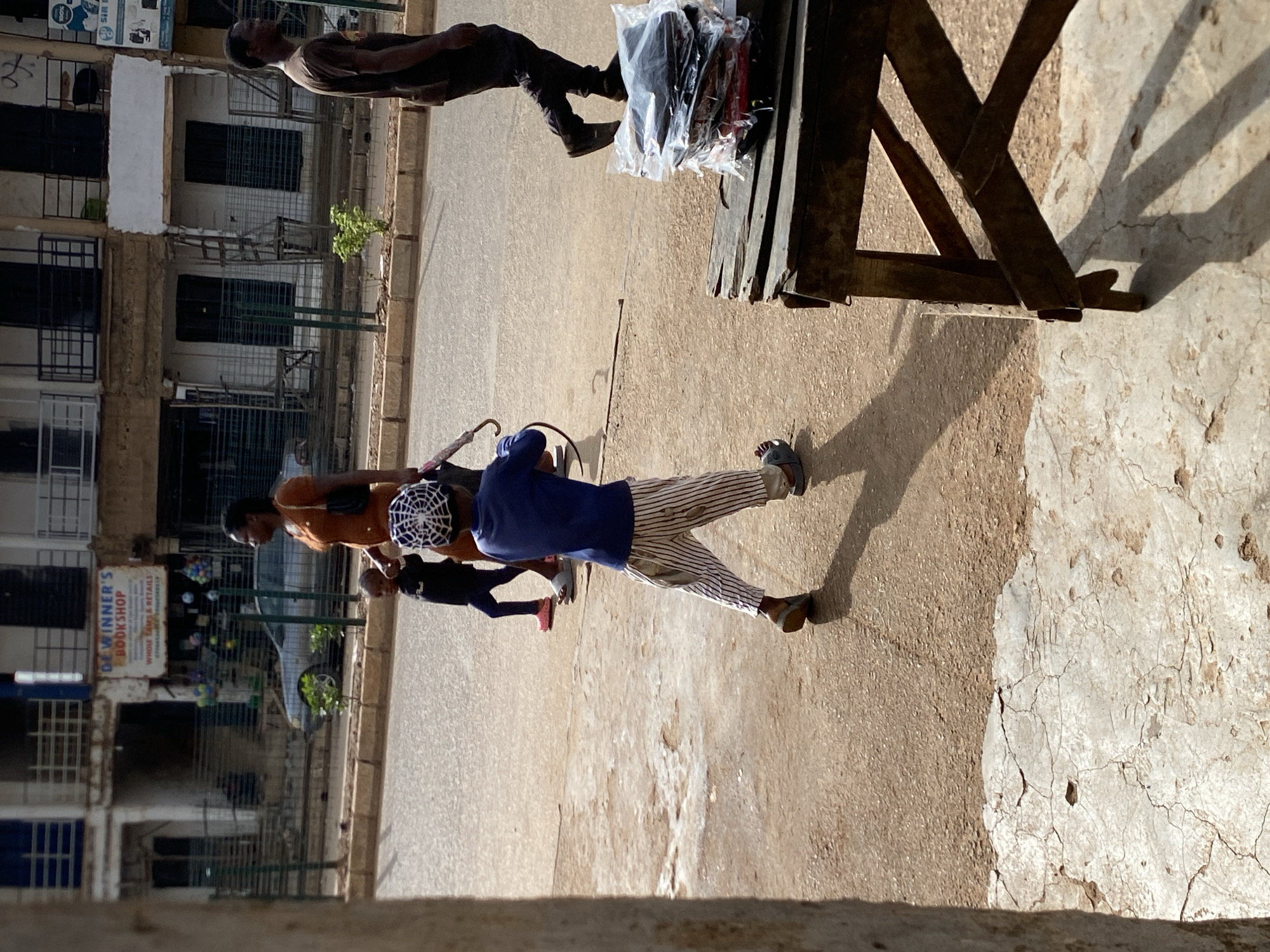 This image captures a bright, sunny street scene, likely in a market or commercial area of Bukuru, Nigeria. The strong sunlight casts distinct shadows from people and structures onto the dusty-looking paved ground, suggesting it is midday or early afternoon.
In the mid-ground, a wooden stall or table is positioned on the right, laden with merchandise, possibly clothing or other goods, in clear plastic bags. A person, wearing a dark short-sleeved top and dark trousers, stands behind the stall, appearing to be a vendor. On the left side of the street, several individuals are present. One person, wearing a blue long-sleeved top, distinctive white and black striped trousers, and a white cap with a spiderweb-like pattern, looks downwards, possibly attending to something just out of frame or looking at the ground. Behind them, a smaller person (a child) in dark clothing and red shoes is partially visible. Further left, another person, wearing an orange top and carrying a dark bag, walks or stands facing the left side of the frame. A dark-colored car is partially visible parked further down the street.
The background features multi-story buildings lining the street. These structures have numerous windows and balconies, many fitted with protective bars or grills. On one building to the left, a sign is clearly visible, reading "DE WINNER'S BOOKSHOP WHOLE SALE & RETAILS," along with a partial phone number starting with "0706." Another sign, further right on a different building, shows "SIR R" alongside icons that appear to represent electronic devices like a printer, laptop, and phone. The overall scene depicts casual daily life and commerce in an urban setting.
