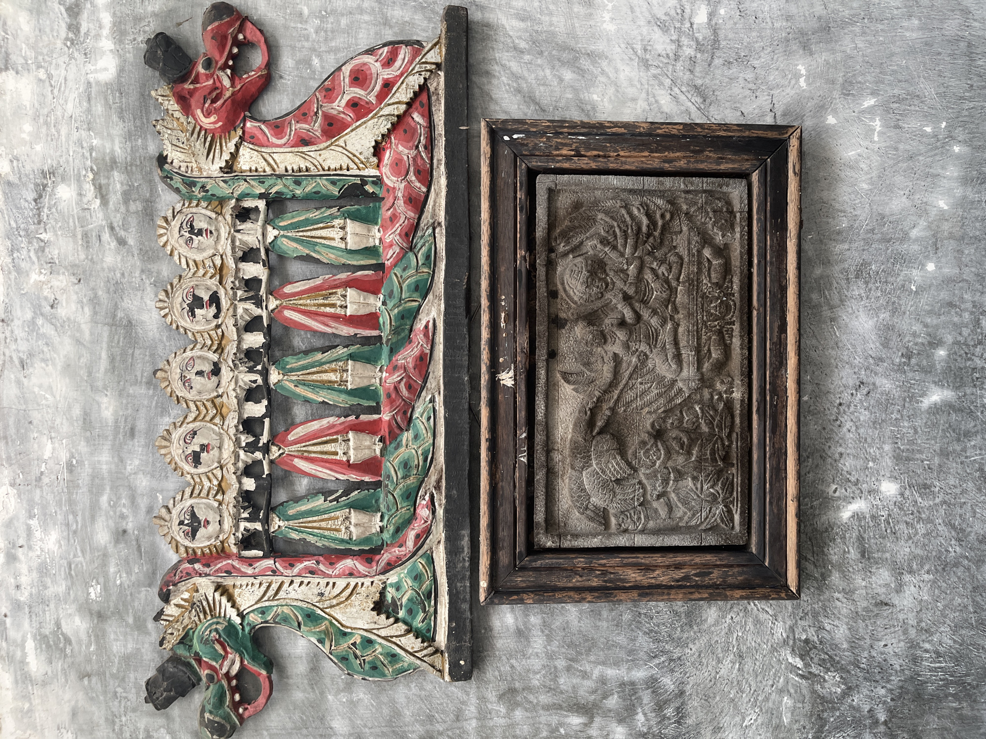 The image shows two pieces of art displayed on a textured gray concrete wall. The upper artwork is a colorful wooden carving featuring two dragon heads on either side, with a row of five figures with distinct facial features and elaborate attire in between. The figures appear to be standing with their arms held down, and their clothing is depicted in red and green with white accents. The lower artwork is a framed stone relief carving. The frame is made of dark, weathered wood. The relief depicts a scene with multiple figures in a stylized, detailed manner, suggesting a narrative or mythological subject. The overall aesthetic suggests traditional Indonesian folk art or cultural artifacts. The location is identified as Karangkebagusan, Indonesia, indicating a specific regional context for these art pieces.