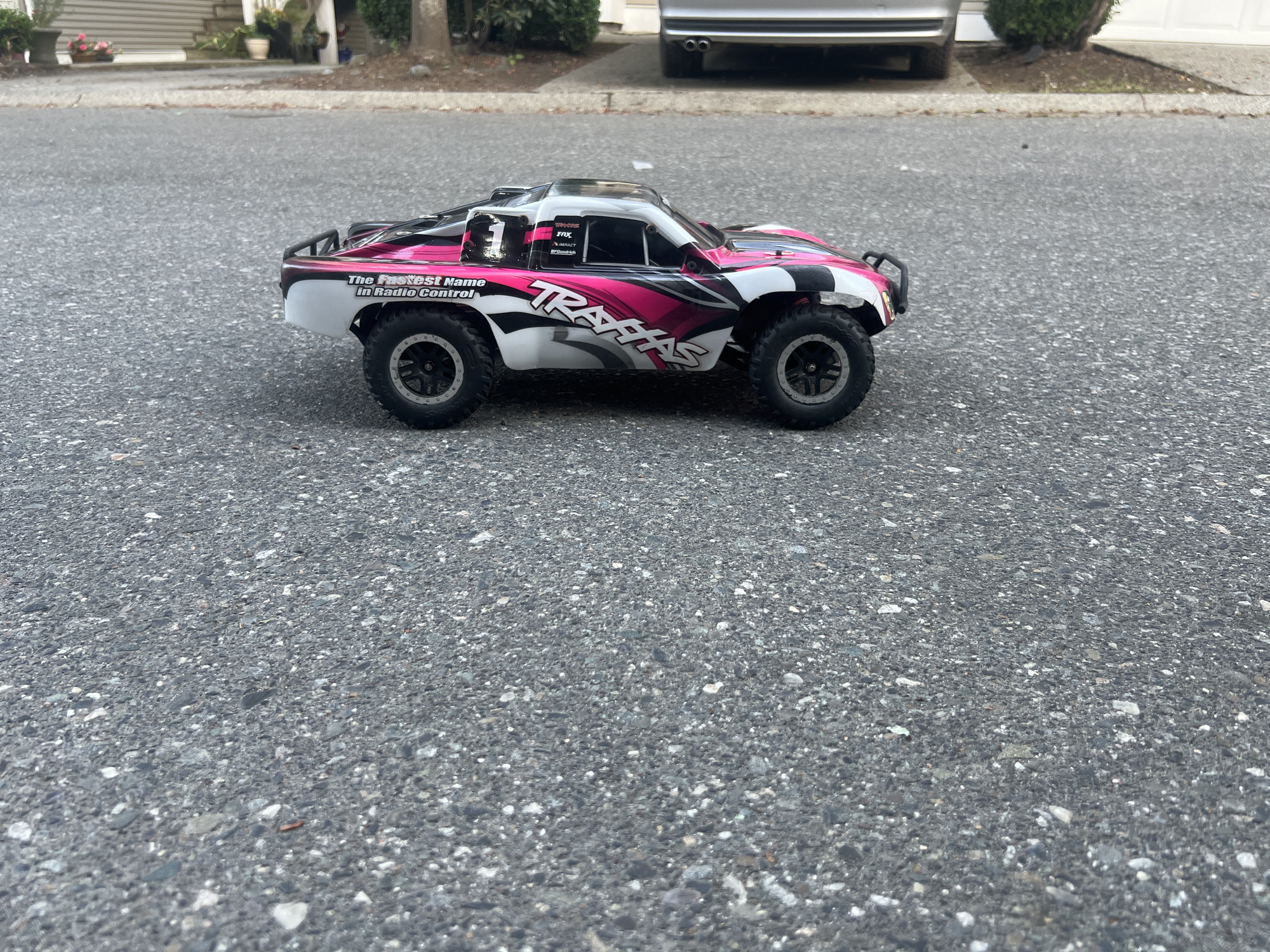 An outdoor, eye-level, medium shot captures a Traxxas radio-controlled monster truck on a paved surface. The truck, predominantly white with pink and black graphics, is positioned in the foreground, facing right. Its wheels are black with silver rims. Behind the truck, a silver car is partially visible, parked on a street. The background features a residential area with houses, trees, and a sidewalk. The overall lighting suggests an overcast day.