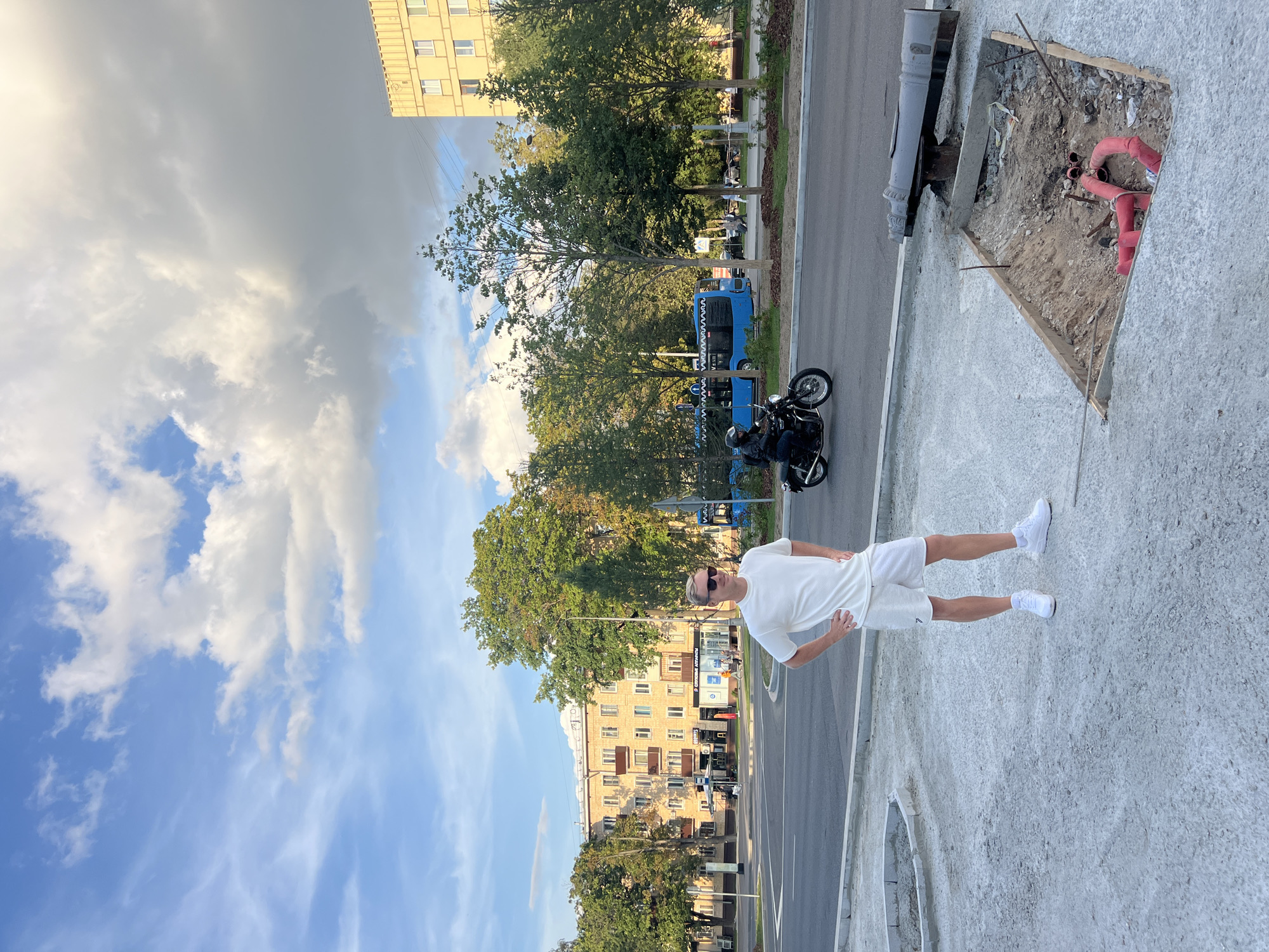A man in a white t-shirt and white shorts stands with his hands on his hips in a city square. He is wearing sunglasses and white sneakers. Behind him, a blue bus drives past on a street, and a motorcycle is also on the street. In the background, there are several trees and buildings, including a yellow apartment building. In the foreground, there is a construction site with a hole in the ground where red pipes are visible. The sky is blue with white clouds. The scene appears to be in Moscow, Russia, given the architecture and the presence of a blue bus which is common public transport in Moscow.