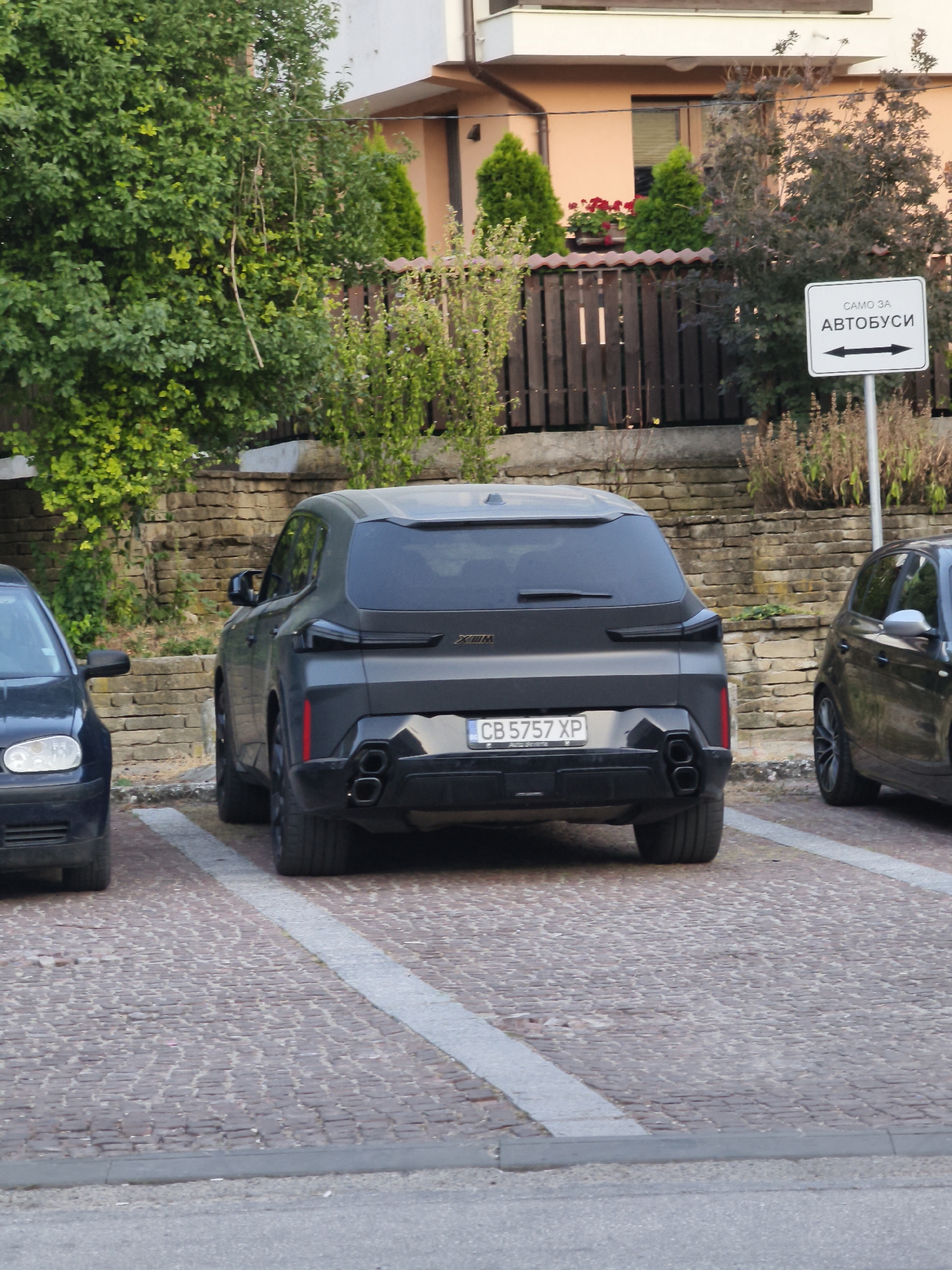 A grey BMW X6 M is parked on a cobblestone surface in Lovech, Bulgaria. The license plate reads "CB 5757 XP". To the left of the BMW, a dark blue car is partially visible. In the background, a wooden fence and a building with a peach-colored facade are present. A white sign with black text and arrows indicating "ONLY FOR BUSES" is mounted on a pole to the right of the BMW. Green foliage is abundant in the scene.