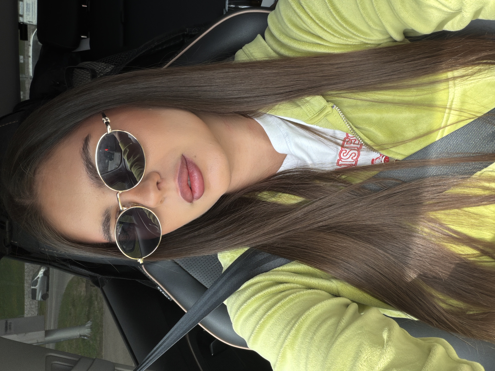 A young woman is in a car, wearing a yellow-green jacket, a white shirt with red letters, and round sunglasses. She has long brown hair. She is wearing a seatbelt. The exterior visible through the car window suggests an urban area, possibly in Krasnoyarsk, Russia.