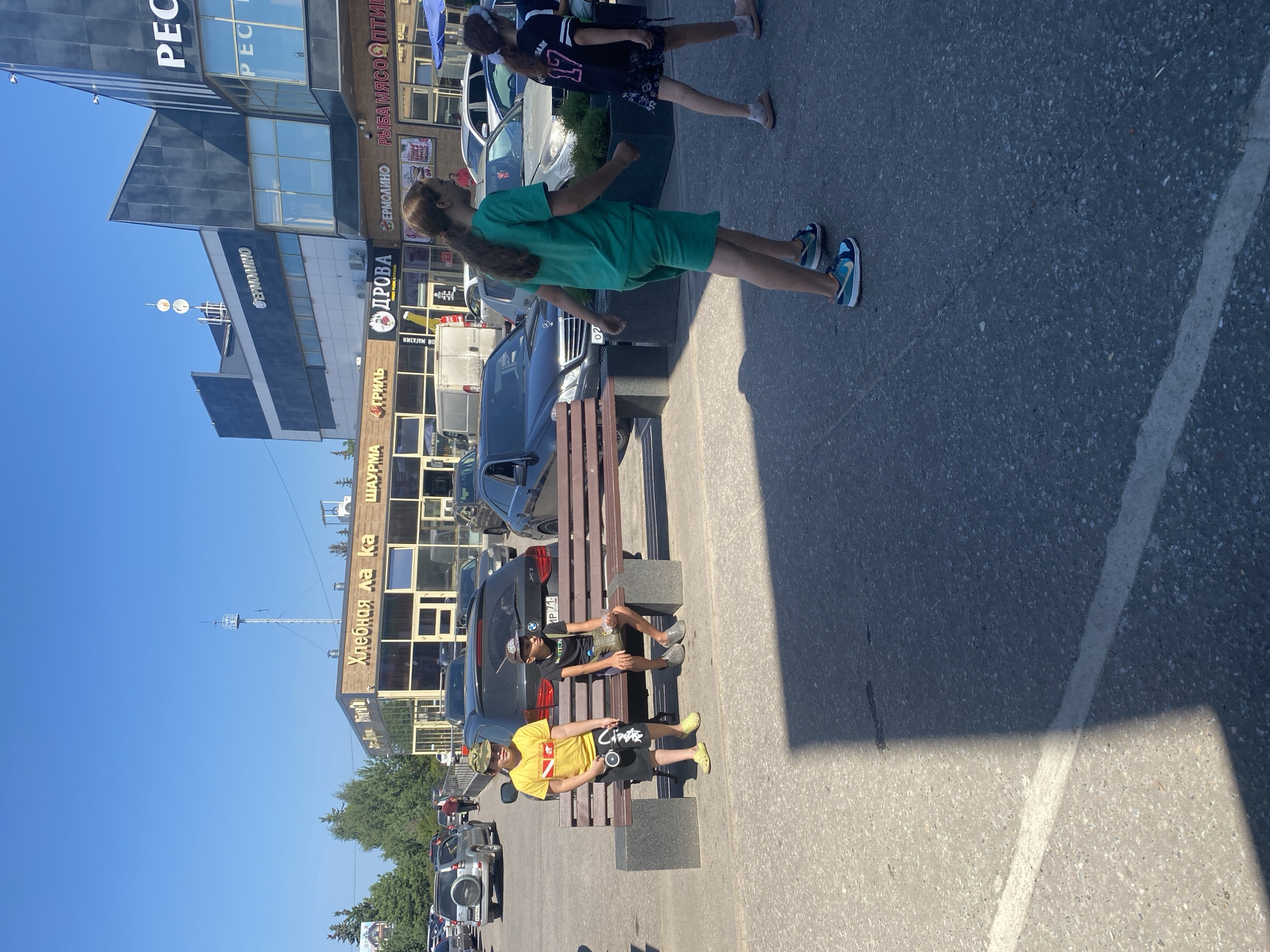 In an outdoor scene in Zaokskiy, Russia, on a clear, sunny day, several people are visible in a parking lot. Two young boys are sitting on a wooden bench. The boy on the left, wearing a yellow t-shirt and black shorts, stands with a toy in his hand. The boy on the right, in a black t-shirt and shorts, sits with a snack. In the background, a building with storefronts is visible, with signage including "ХЛЕБНАЯ ЛАКА," "ШАУРМА," "ГРИЛЬ," "ДРОВА," and "ЕРМОЛИНО." Several cars are parked in the lot, and a few people are walking in the vicinity. A young girl in a green outfit walks towards the right of the frame, and another girl in a black shirt and patterned skirt is seen further to the right, walking away. The environment appears to be a commercial area with a paved parking lot and a clear blue sky overhead.
