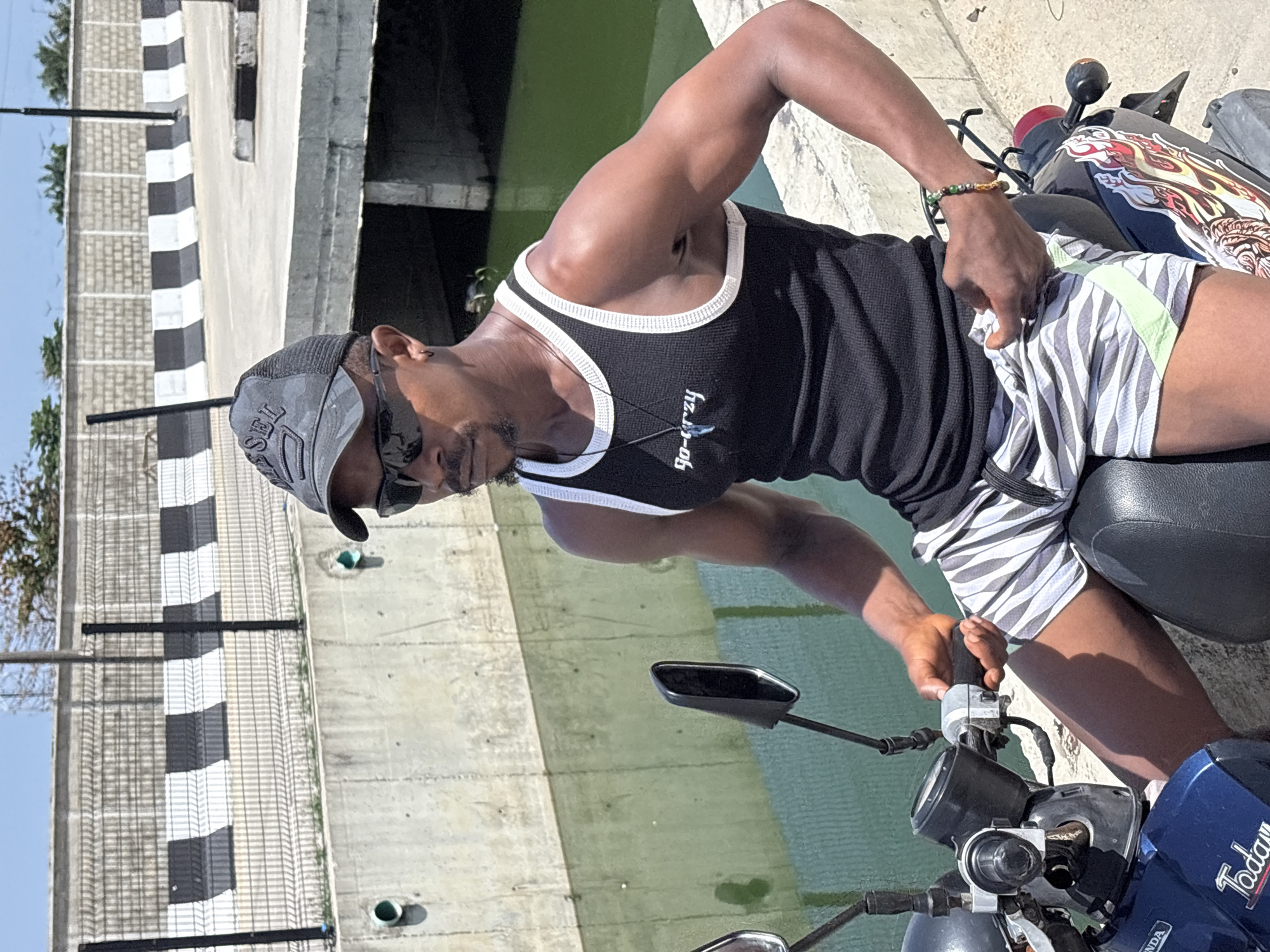 A person is seated on a motorcycle in Igboefon, Nigeria, near a concrete structure with black and white barriers. The individual, wearing a black tank top with white trim, striped shorts, a baseball cap, and sunglasses, is holding the motorcycle's handlebars. The motorcycle has a blue front panel and a design on the seat. The background features a body of water.